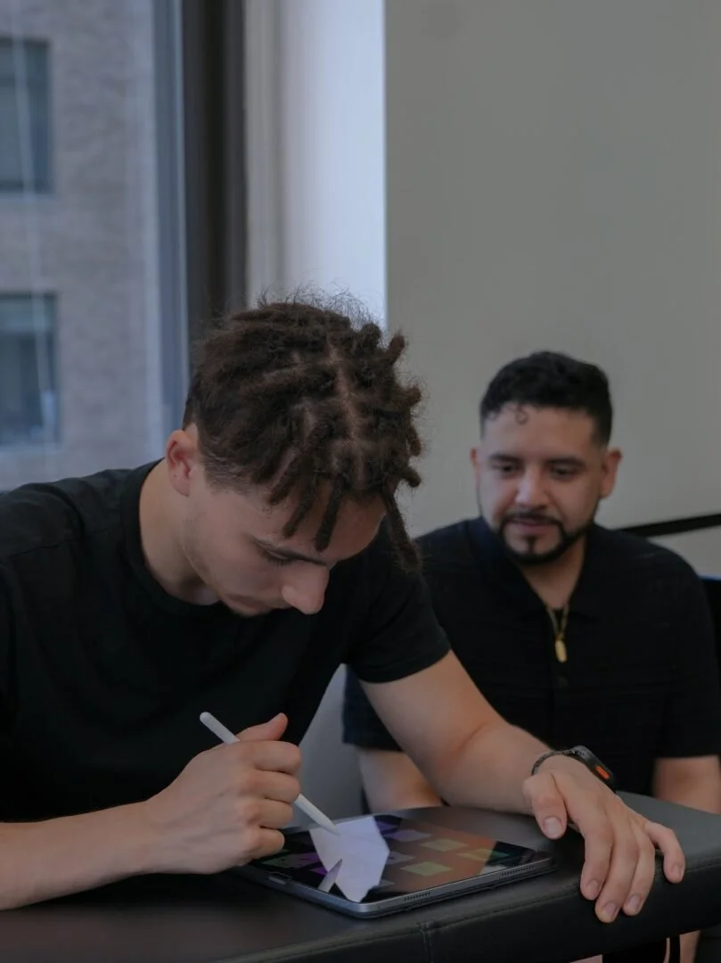 Two men sitting at a table, one drawing on a tablet with a stylus, and the other watching.