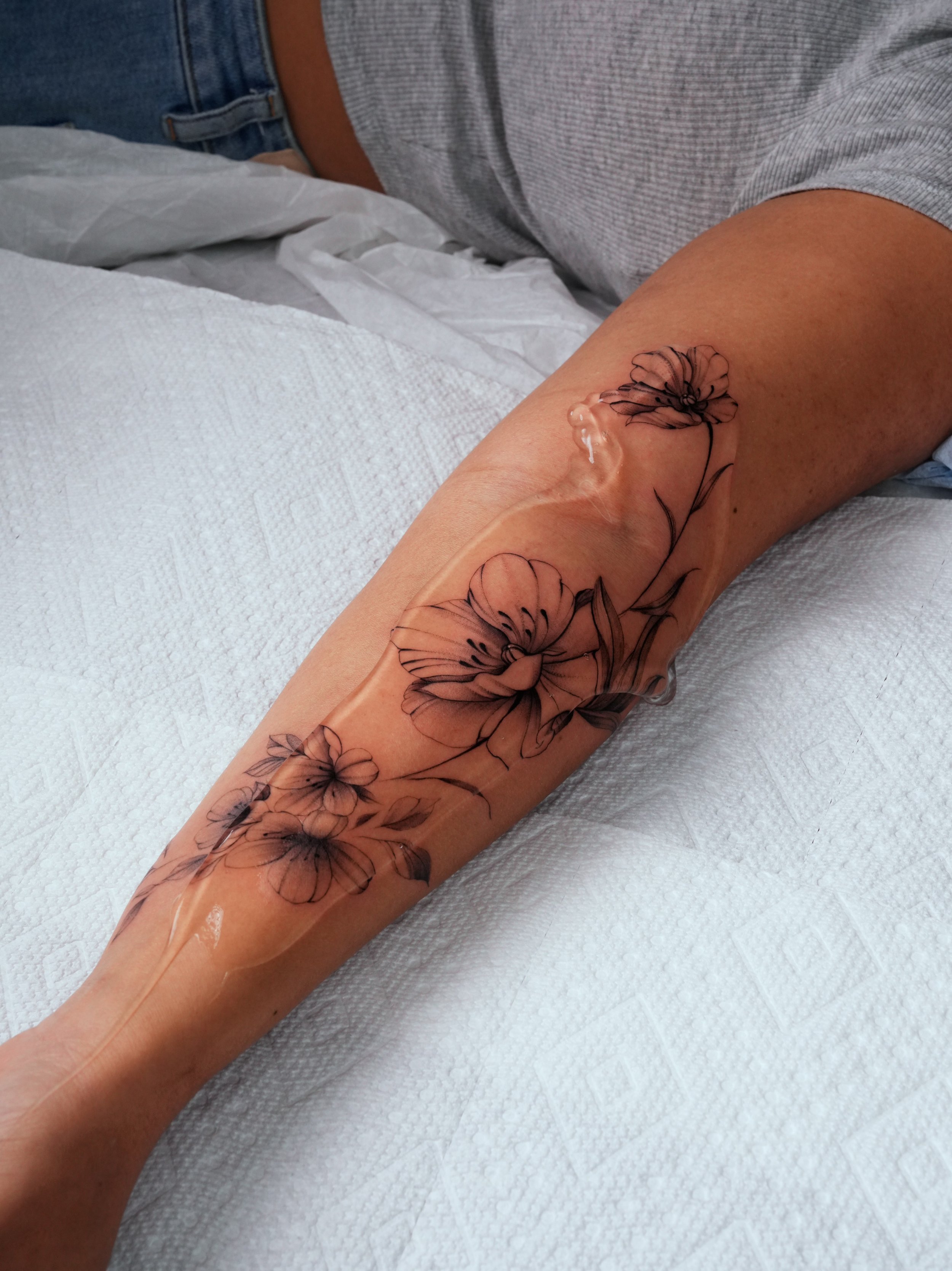Close-up of a person's arm with a floral tattoo, lying on a white surface, with part of their torso visible wearing a gray shirt and jeans.