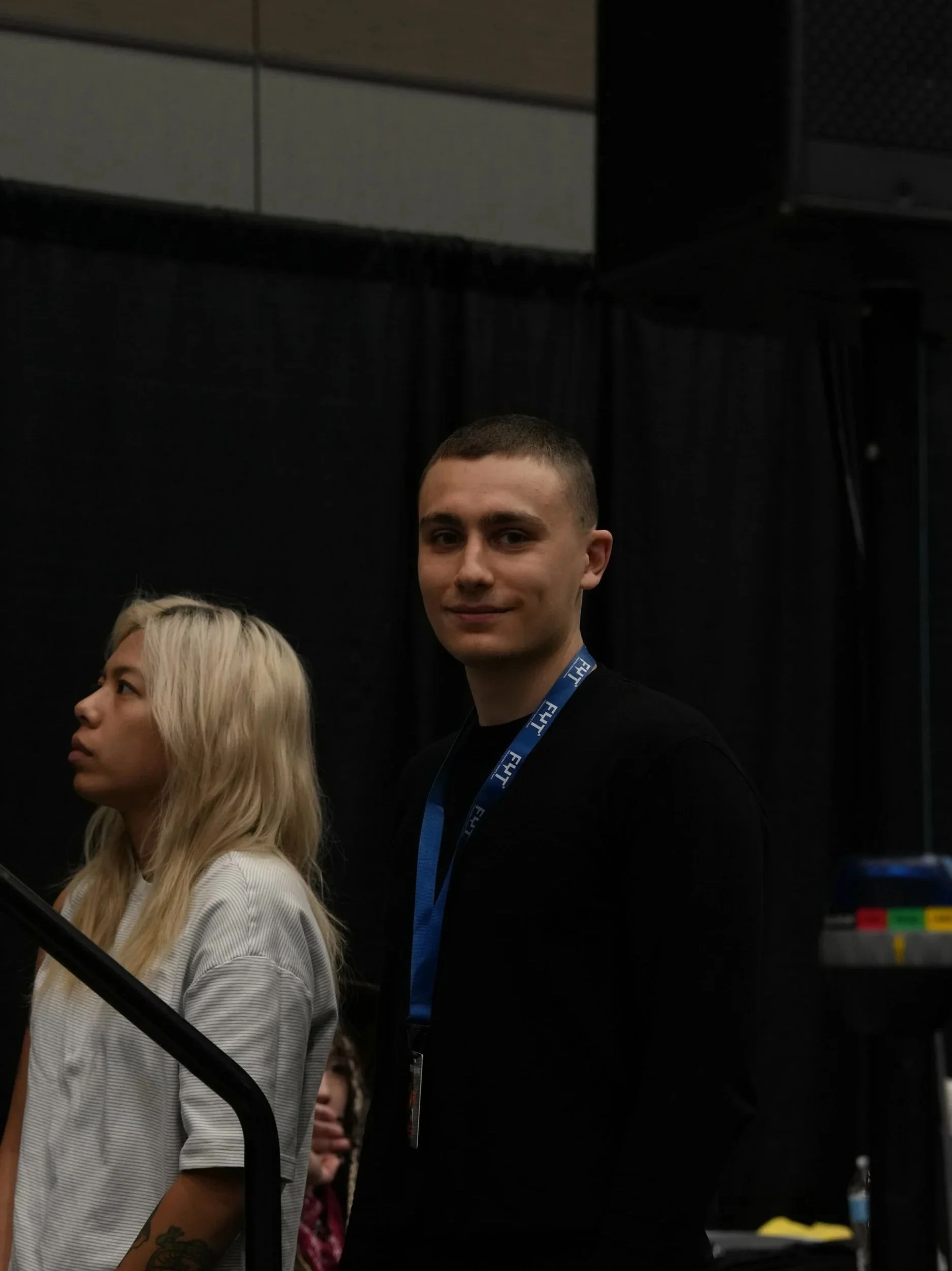 A young man with short brown hair and a blue lanyard smiling, standing next to a blonde woman with a tattoo on her arm and a striped shirt, in a dark indoor setting with a black background and some machinery or equipment visible.
