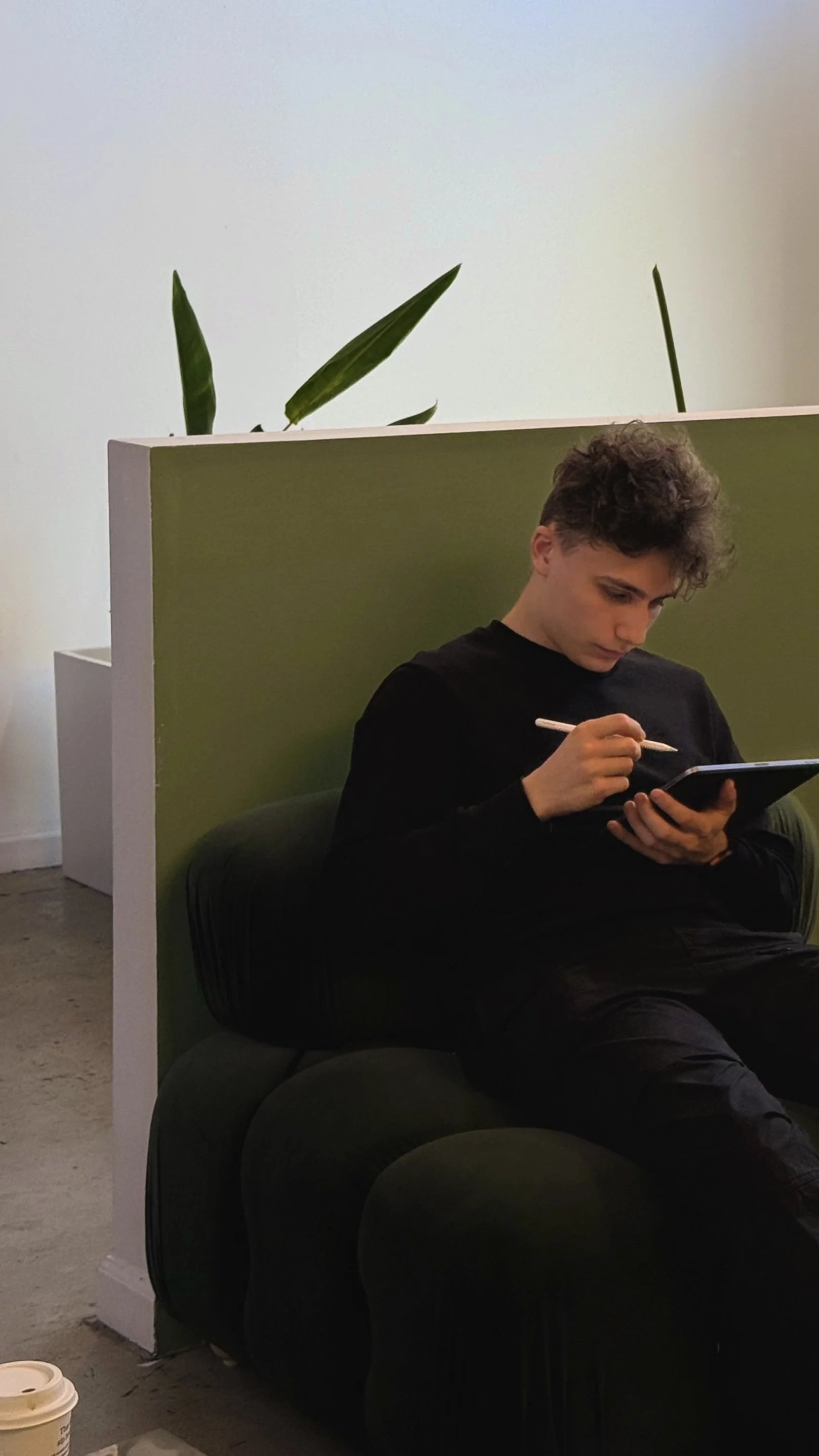 A young man with curly hair wearing a black shirt sitting on a green chair, holding a notebook and pen, partially behind a green partition, with a potted plant visible behind the partition.
