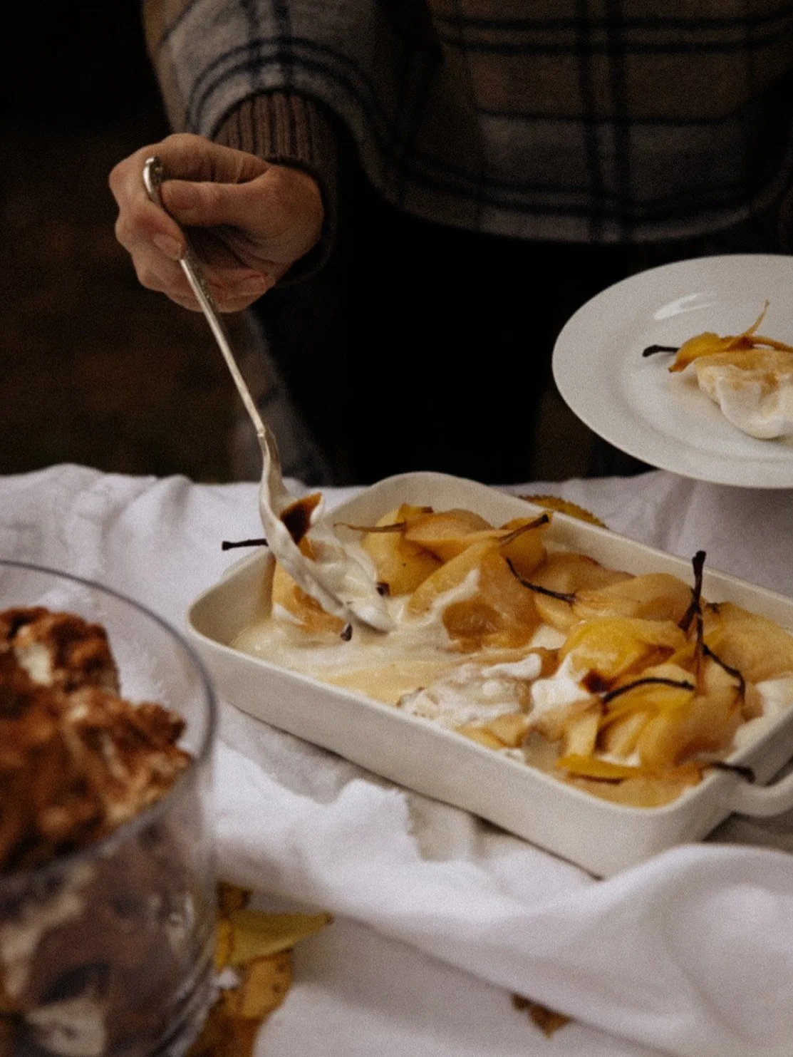 Person serving baked pears in a creamy sauce at a dinner table, with other dishes present.