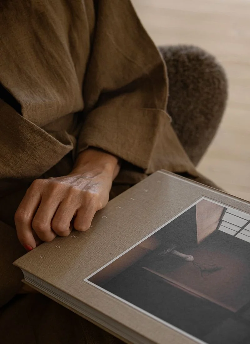 Close-up of a person's hand resting on an open photo book, with a brown sofa and a furry ottoman in the background.