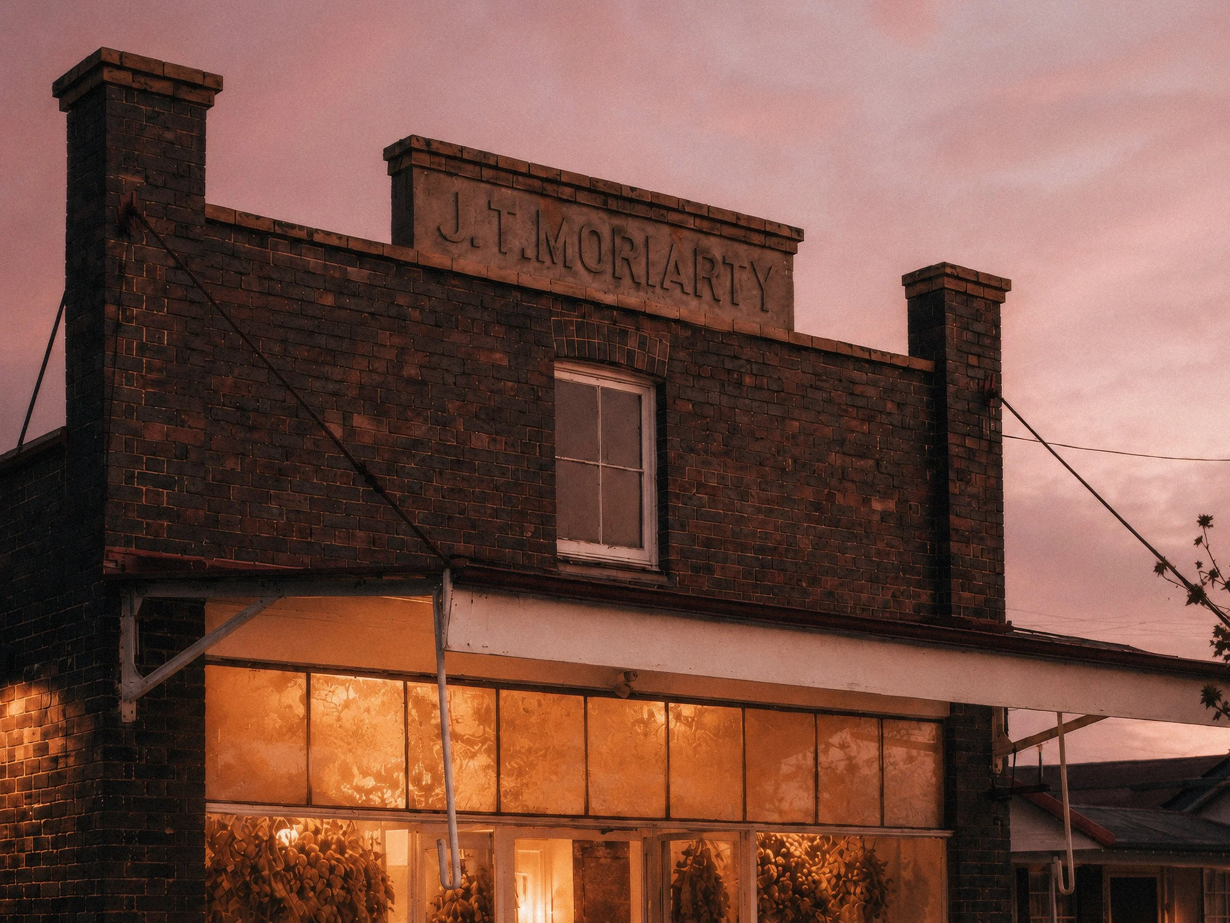 A brick building with a sign that reads 'J. L. MORLARTY' on the top. The building has a window and a glass storefront with warm lighting inside. The sky is a pinkish hue, suggesting it is either dawn or dusk.
