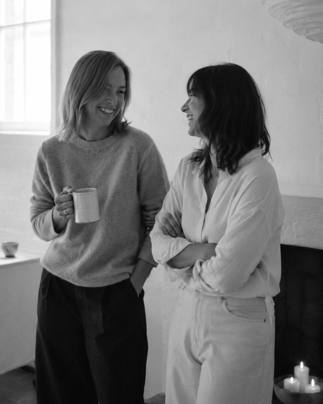 Two women smiling and talking with each other in a cozy room, one holding a mug, with candles on a table in the background.