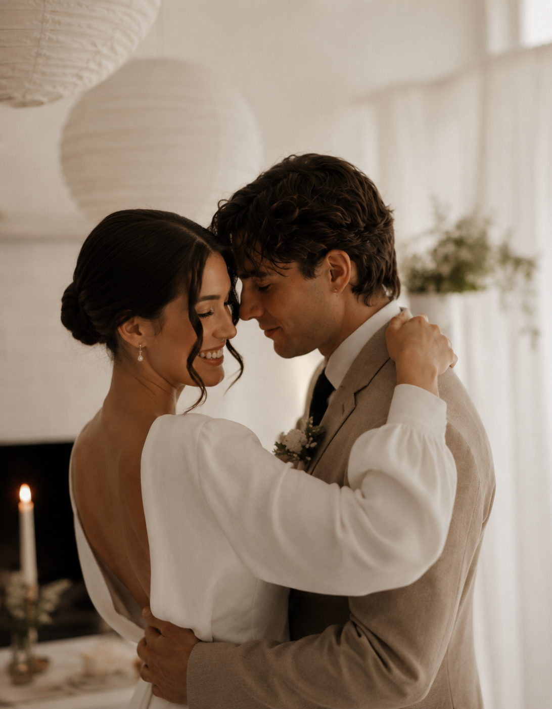 A couple at their wedding dancing closely with foreheads touching, smiling, and eyes closed outdoors, with warm lighting and candles in the background.