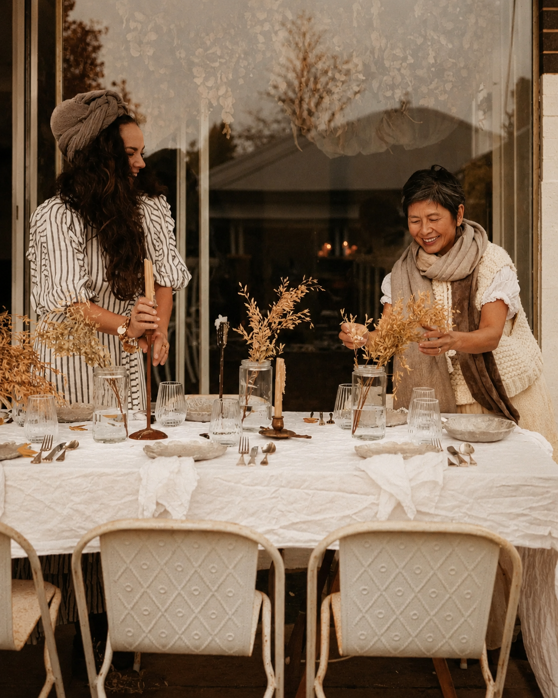 Two women setting a table outdoors with vases of dried branches, glassware, and tableware, smiling and engaging in conversation.