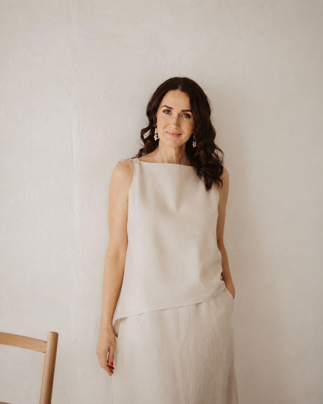 Woman with wavy dark hair wearing a cream sleeveless dress and chandelier earrings, standing against a plain off-white wall.