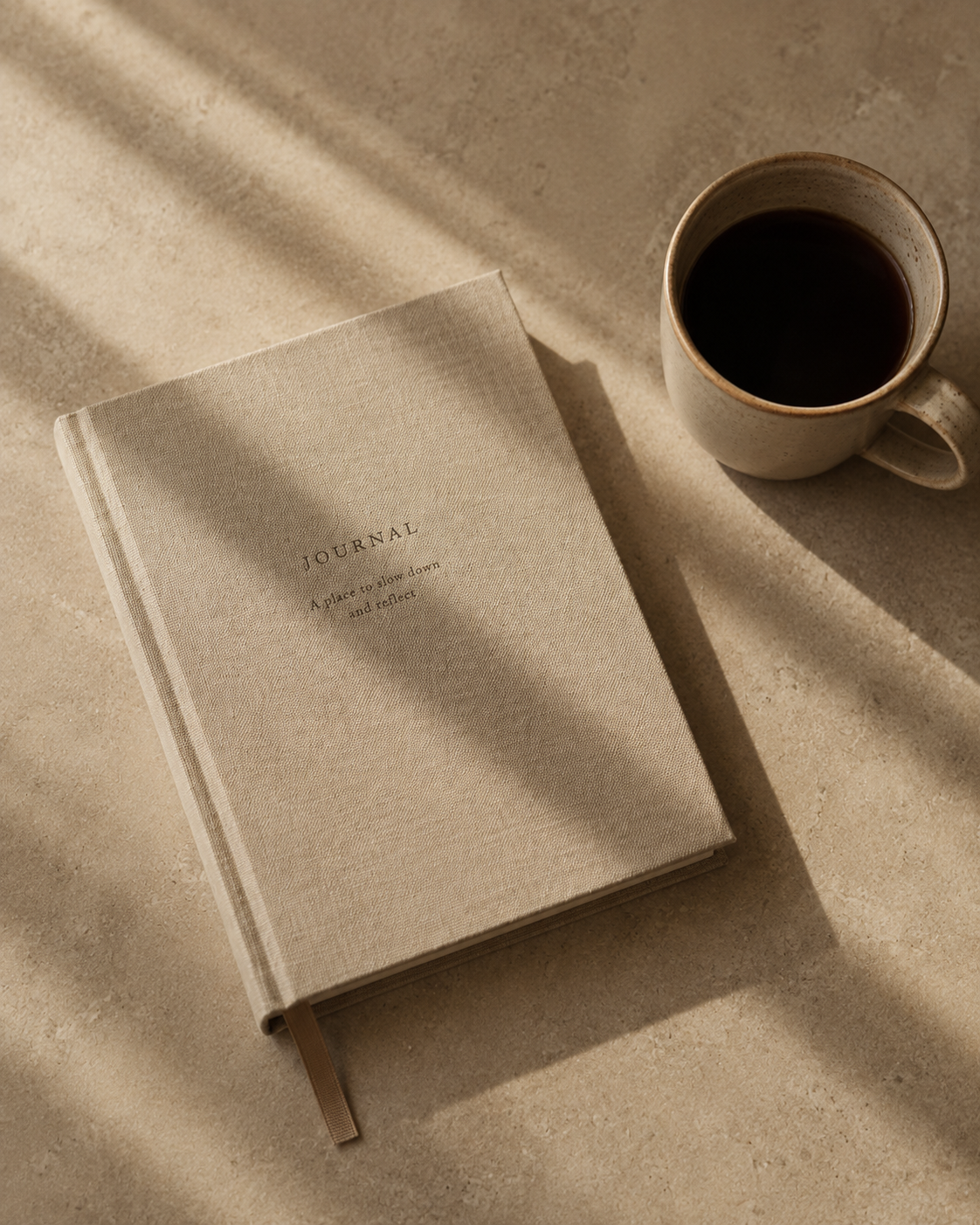 A beige journal with the words 'JOURNAL A place to slow down and reflect' on the cover, placed on a light-colored surface with shadows, next to a mug of black coffee.