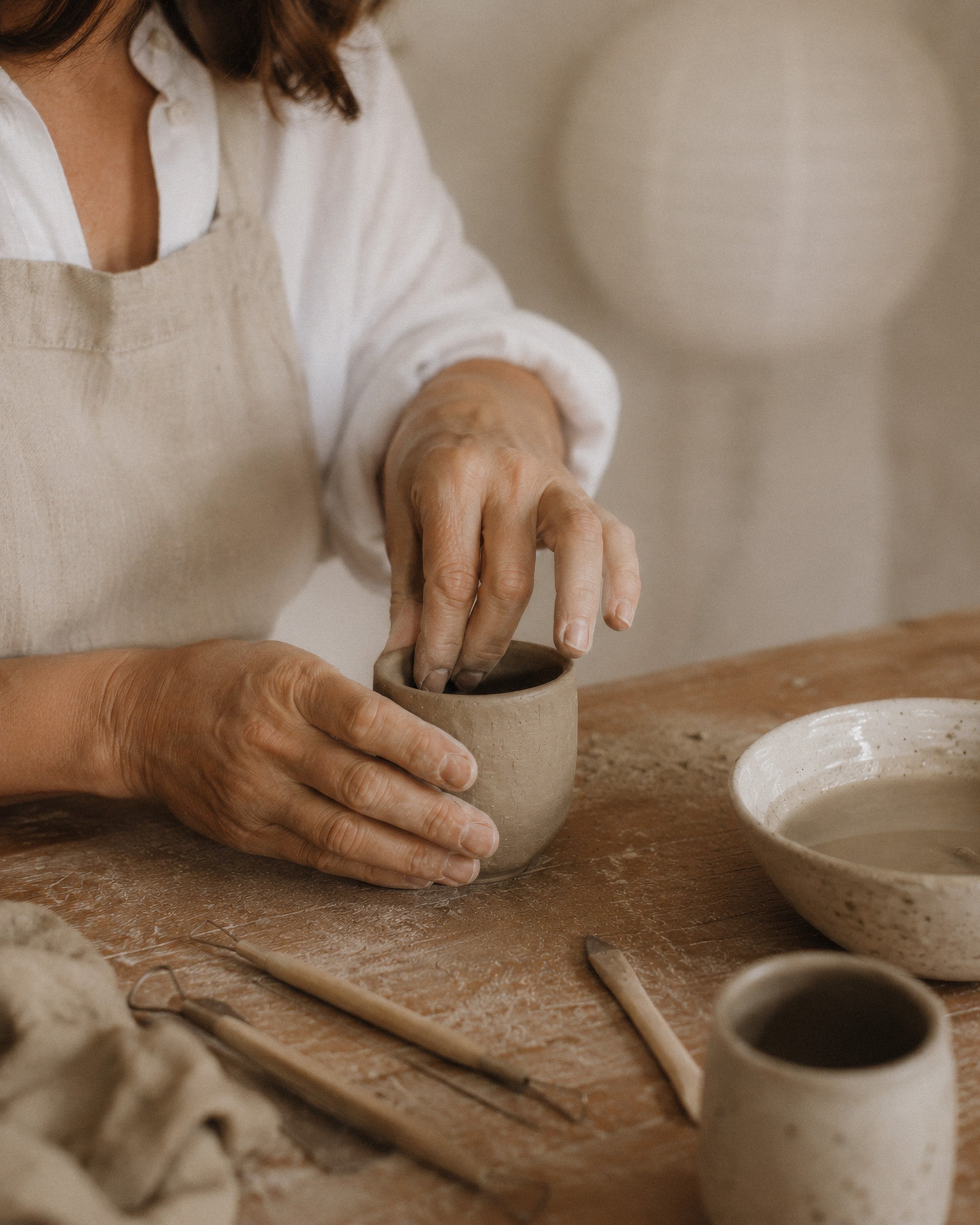Close-up of a woman's hands shaping clay in a small bowl during pottery making, with pottery tools and bowls on a wooden table covered in clay dust.