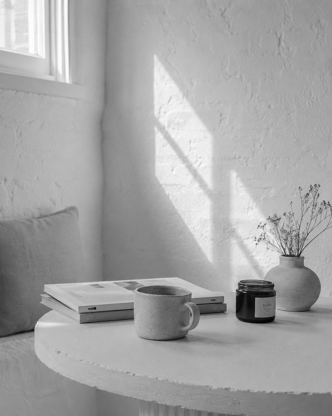 Black and white image of a cozy nook with a round table holding a mug, a jar with a label, and a stack of magazines. A potted plant with delicate branches is on the table, and sunlight creates geometric shadows on the wall.