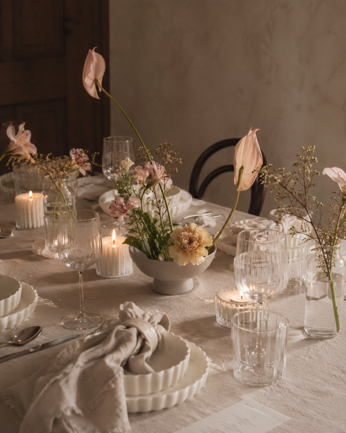 A formal dining table decorated with a beige tablecloth, floral centerpiece with pink lilies and other flowers, lit candles in glass holders, white bowls, and clear glassware, set for a meal.