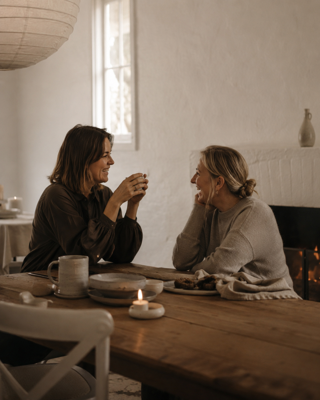 Two women sitting at a dining table, smiling and talking, with a lit candle and plates of food.