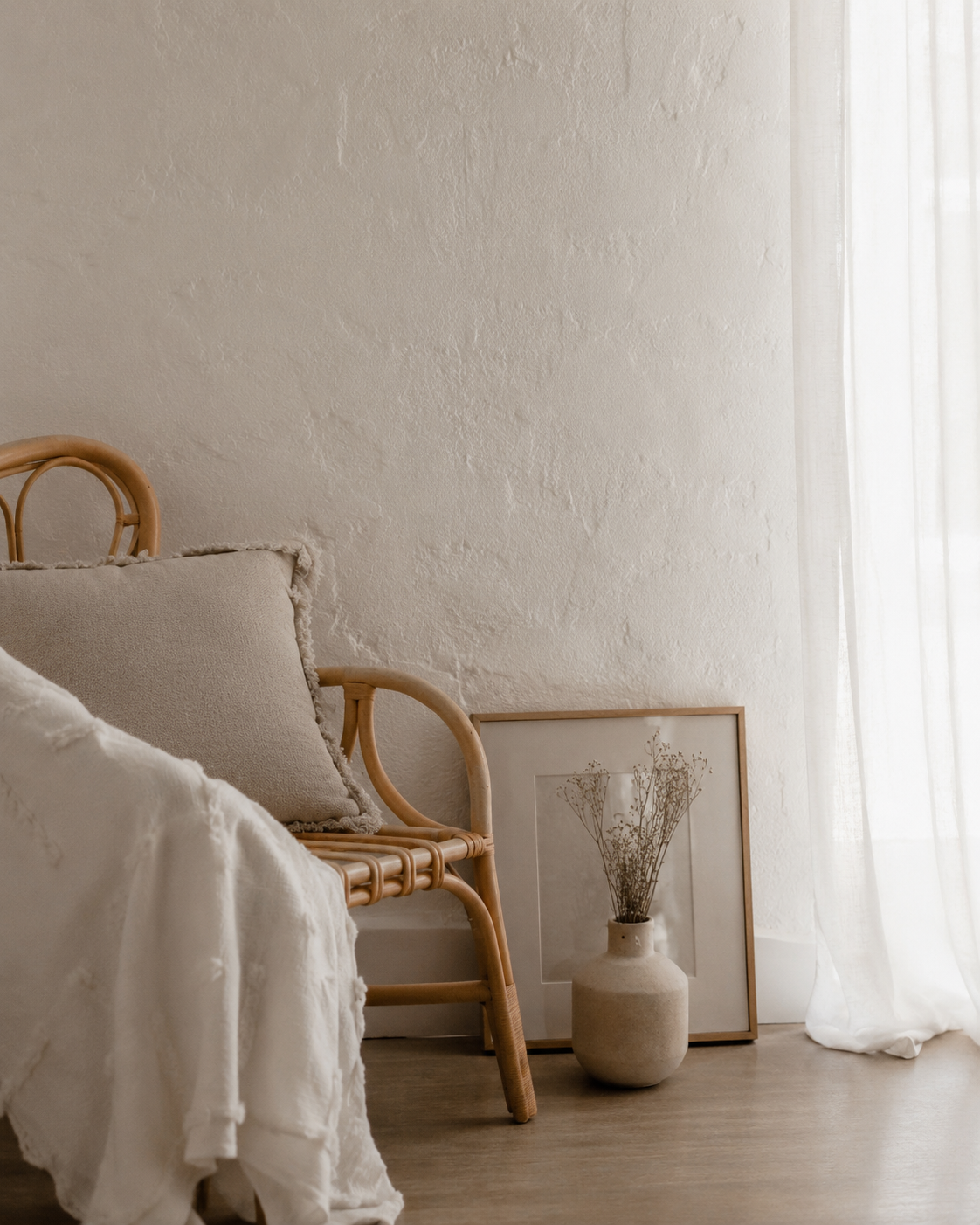 A cozy bedroom corner featuring a rattan bed with neutral-colored pillows and a blanket, next to a framed dried flower art print leaning against the wall, beside a ceramic vase with dried flowers, and white curtains allowing soft natural light.