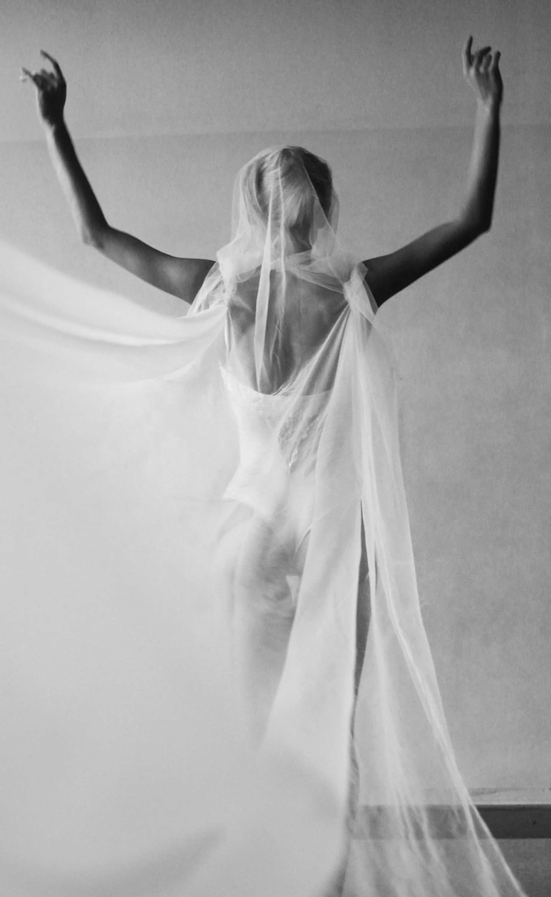 A woman with her back to the camera, wearing a sheer, flowing veil or fabric over her shoulders and head, with her arms raised above her head, in black and white.