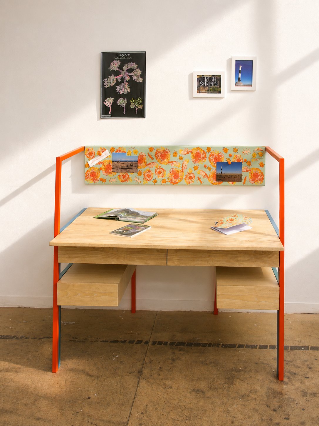 A modern desk with a light wood top and storage drawers, orange metal frame, and a floral backrest. The wall above has framed pictures and a poster, with items pinned to the backrest and papers and magazines on the desk.