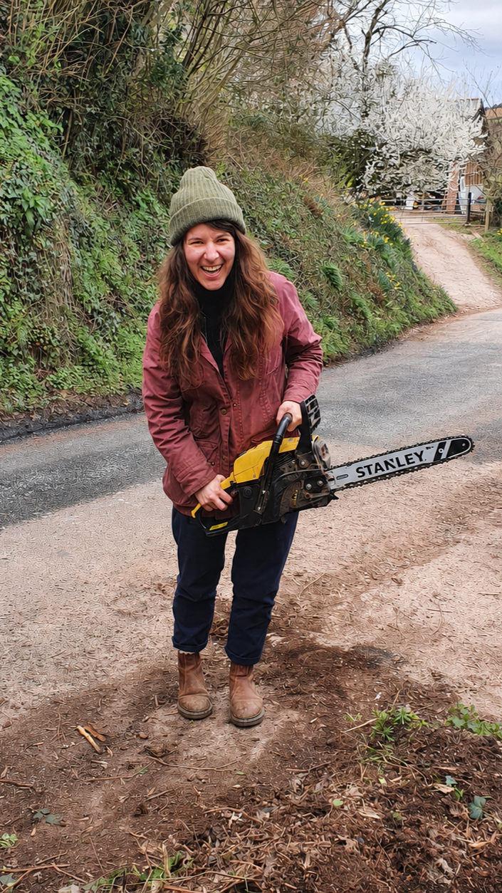 A cheerful woman outdoors holding a yellow and black chainsaw, wearing a green beanie, maroon jacket, black pants, and brown boots, standing on a dirt path with greenery and a white flowering tree in the background.