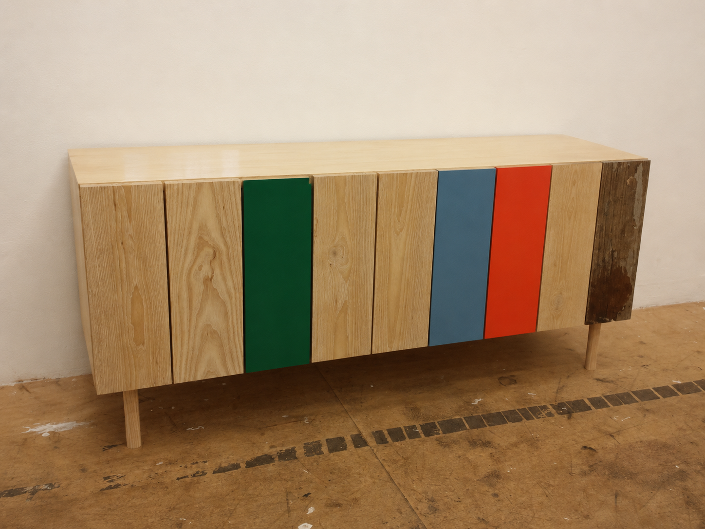 Wooden sideboard with multicolored panels in green, blue, orange, and natural wood, standing against a white wall on a wooden floor.