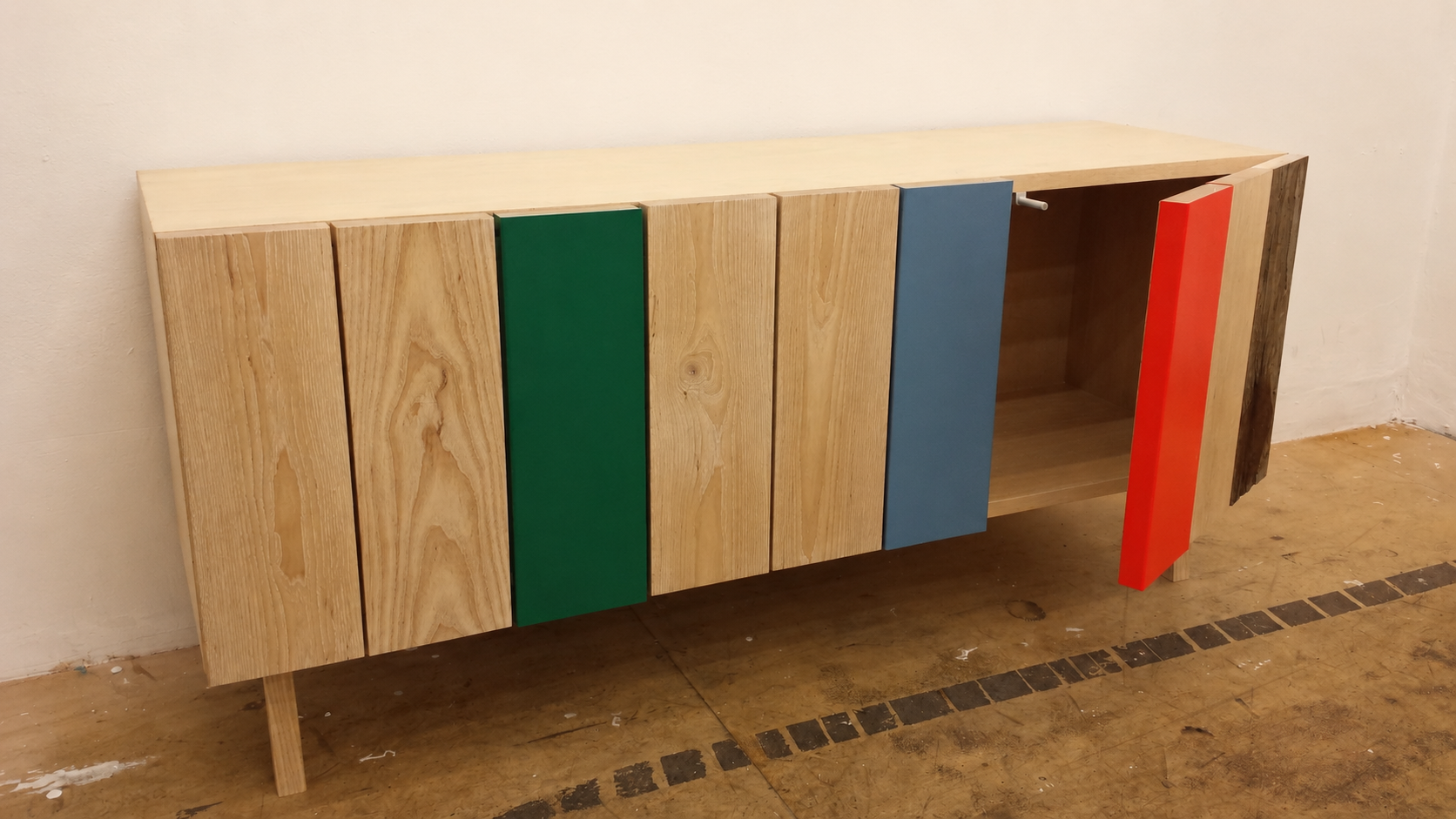 A wooden sideboard with colorful cabinet doors in green, blue, and red, and an open shelf, placed against a white wall with a wooden floor.