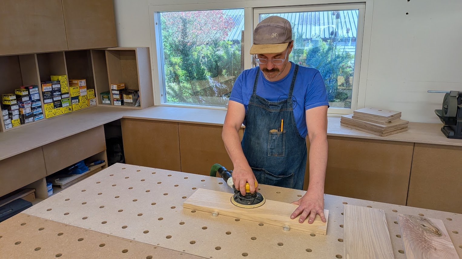 A man with glasses, a blue t-shirt, apron, and a beige cap is sanding a piece of wood in a woodworking shop.