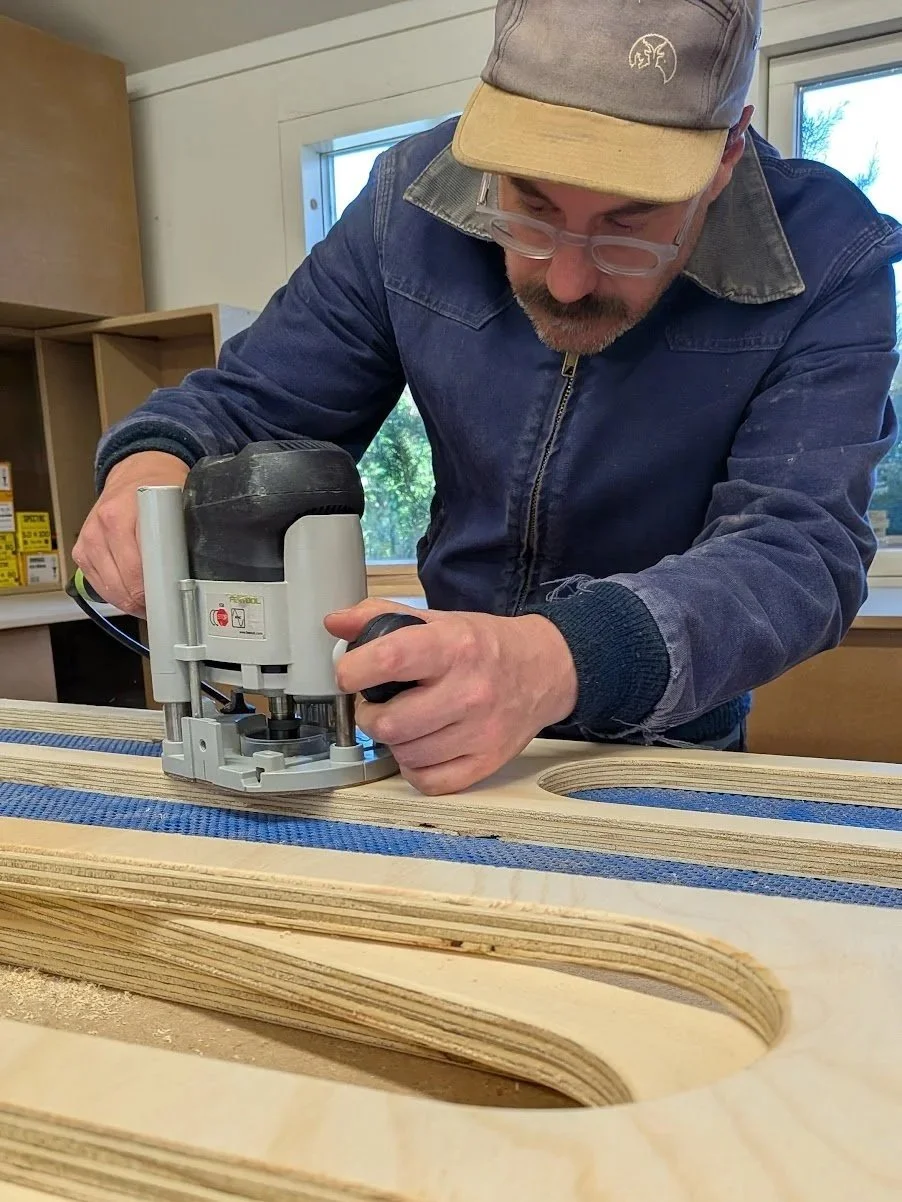 A man wearing glasses, a cap, and a denim jacket is using a router on a piece of bent plywood in a workshop.