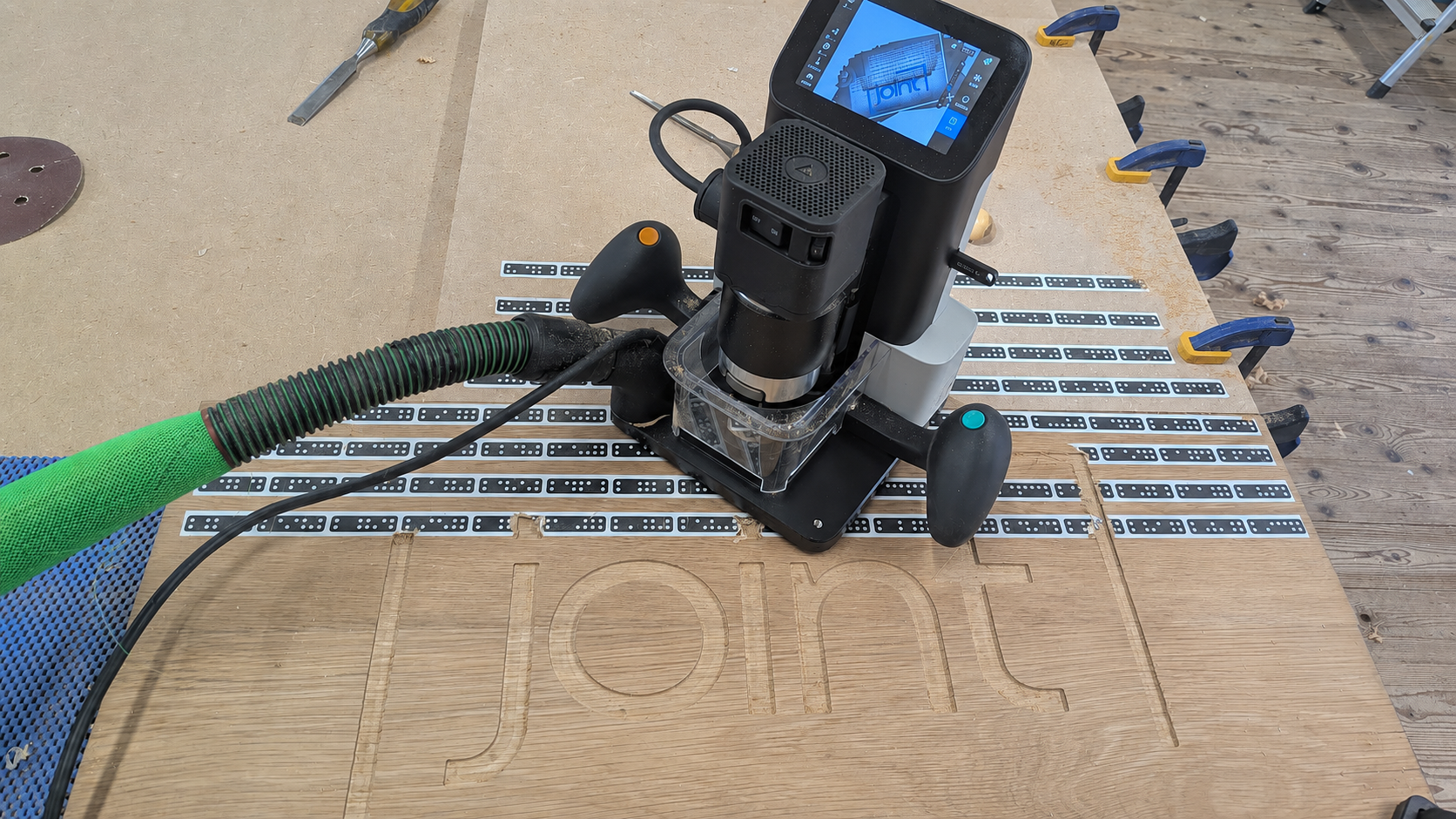A router bit attached to a CNC machine carving the word 'join' into a wooden surface, with various clamps, tools, and a display screen visible.
