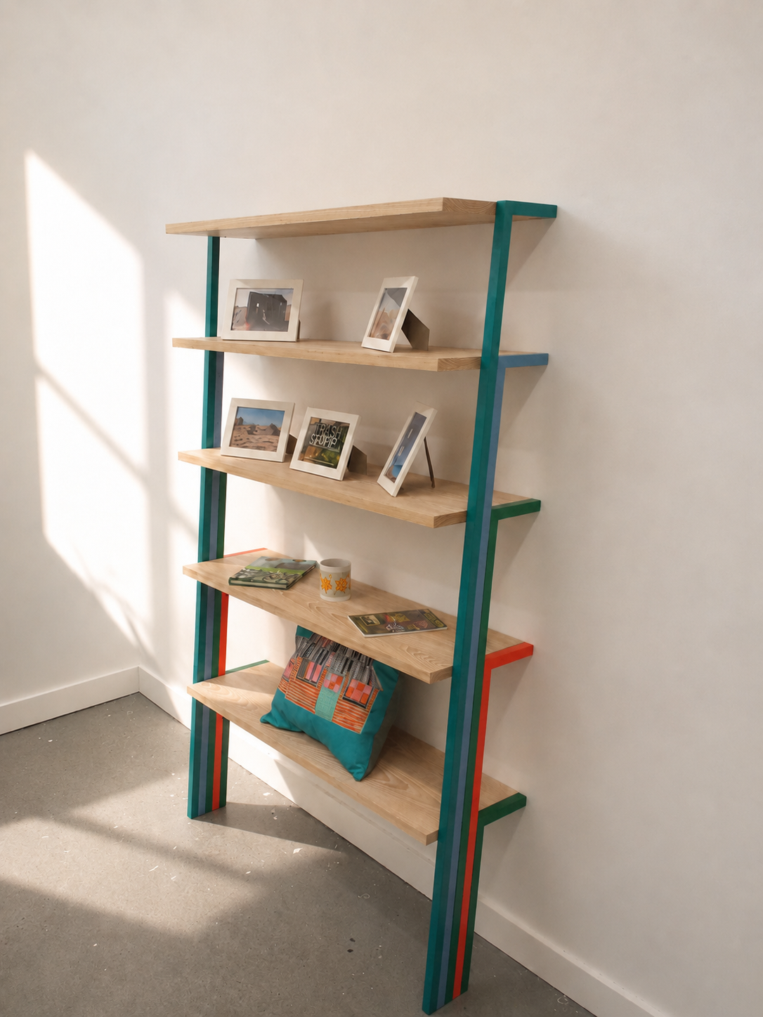 A wooden bookshelf with multicolored sides, holding framed photos, magazines, a mug, and a pillow.