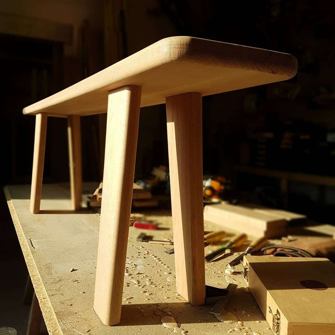 A small wooden stool with a rectangular top and angled legs is placed on a workbench in a woodworking shop.