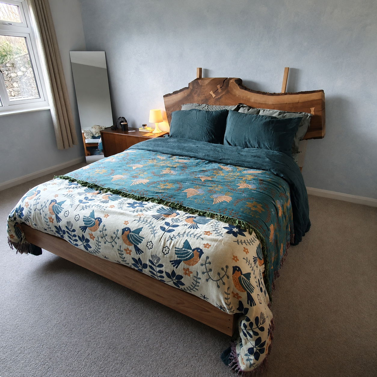 A bedroom with a bed featuring a bird and foliage patterned quilt, dark green pillows, a wooden headboard, a nightstand with a lamp, and a mirror near a window with beige curtains.