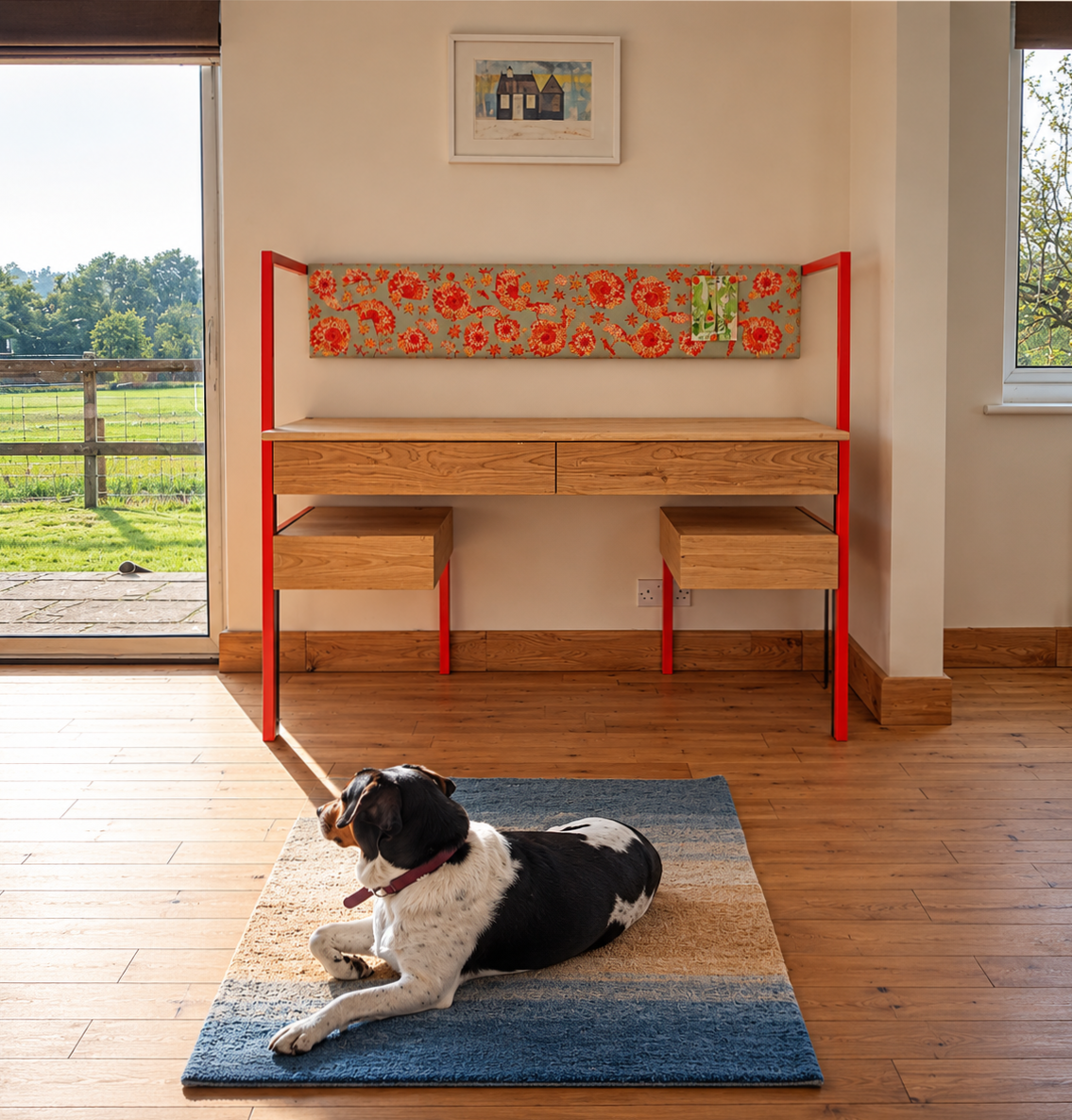 A black and white dog lying on a multi-colored area rug in front of a wooden desk with a red metal frame, inside a room with hardwood floors and beige walls, with windows showing an outdoor view of a grassy yard and trees.