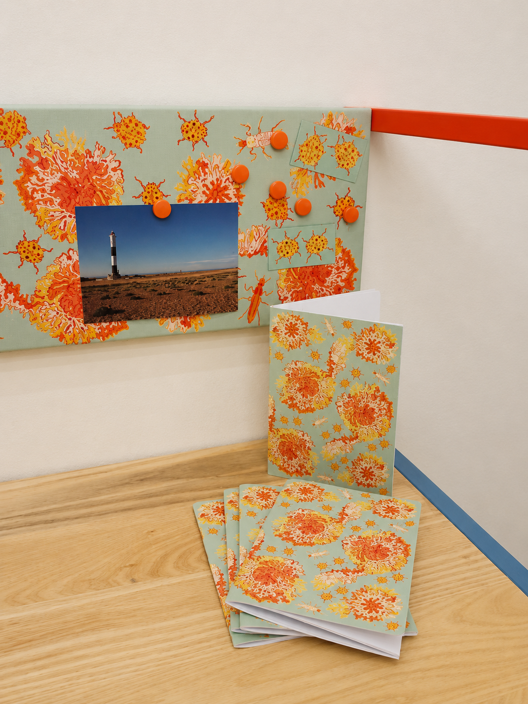 A corkboard decorated with orange and yellow floral-patterned fabric, holding a photo of a lighthouse on a grassy landscape, with several orange magnets and small printed images of orange and yellow creatures. Matching folders and papers with floral patterns are arranged on a wooden surface below.