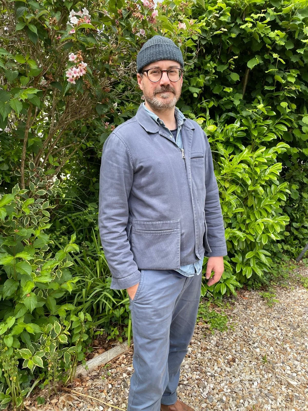 A man wearing glasses, a gray beanie, a gray jacket, and gray pants stands outdoors with hands in pockets, surrounded by lush green foliage and pink flowers.