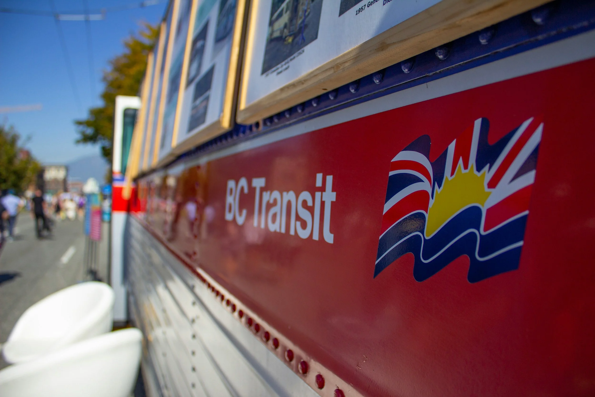 Close-up of a red BC Transit bus with advertisements, with a blurred background of trees and people.