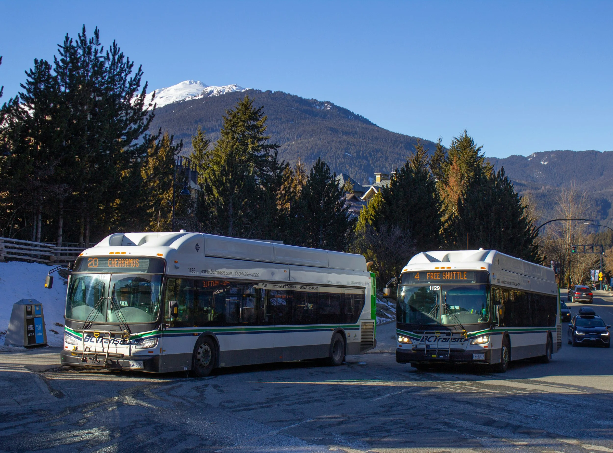 Two BC Transit buses parked at a bus stop with snow on the ground, mountains with snow in the background, and trees surrounding the area.