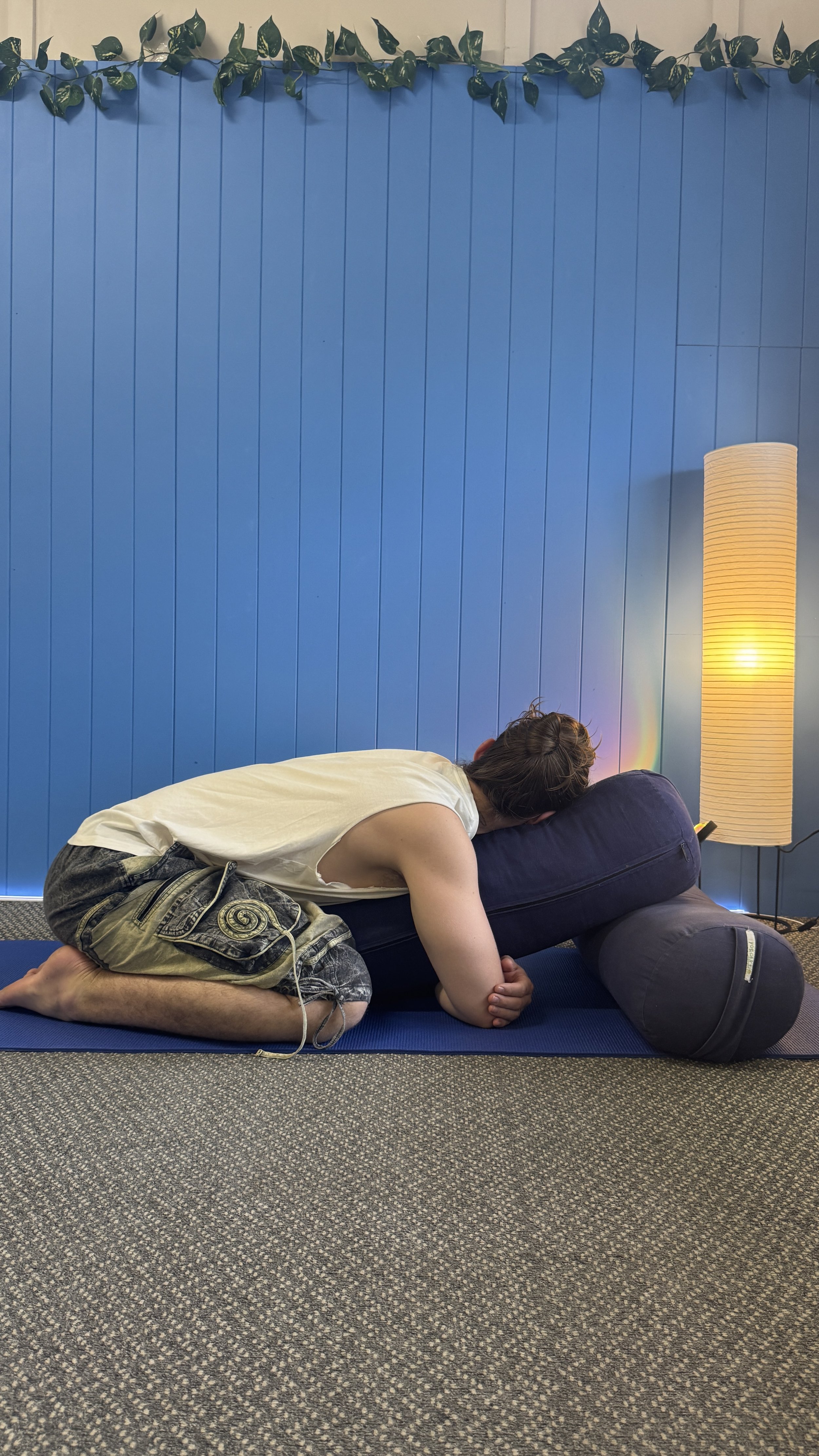 A person with short hair and wearing casual clothes is doing a yoga pose on a blue mat indoors. They are kneeling and leaning forward with their head resting on a pillow. There is a blue wall with greenery at the top, a tall white lamp, and a carpeted floor.