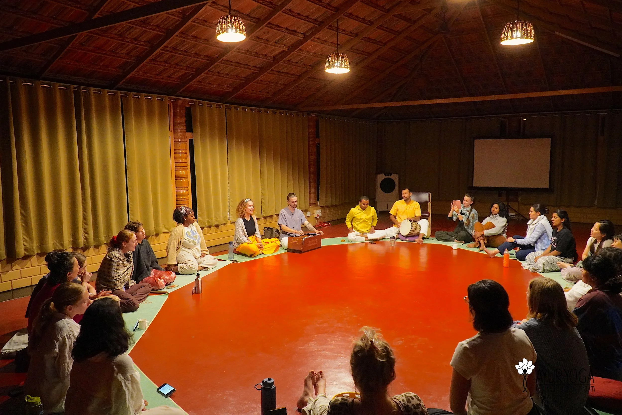 A group of people sitting in a circle on the floor in a room with yellow curtains and wooden ceiling, participating in a meditation or spiritual practice, with some holding musical instruments and others sitting peacefully.