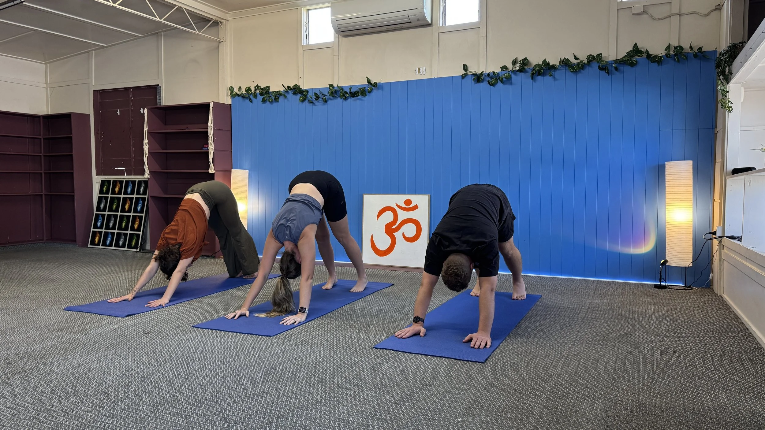 Three people practicing yoga in downward dog pose on blue mats in a studio with purple and blue walls, decorated with a plant garland and a sign with an orange Om symbol.