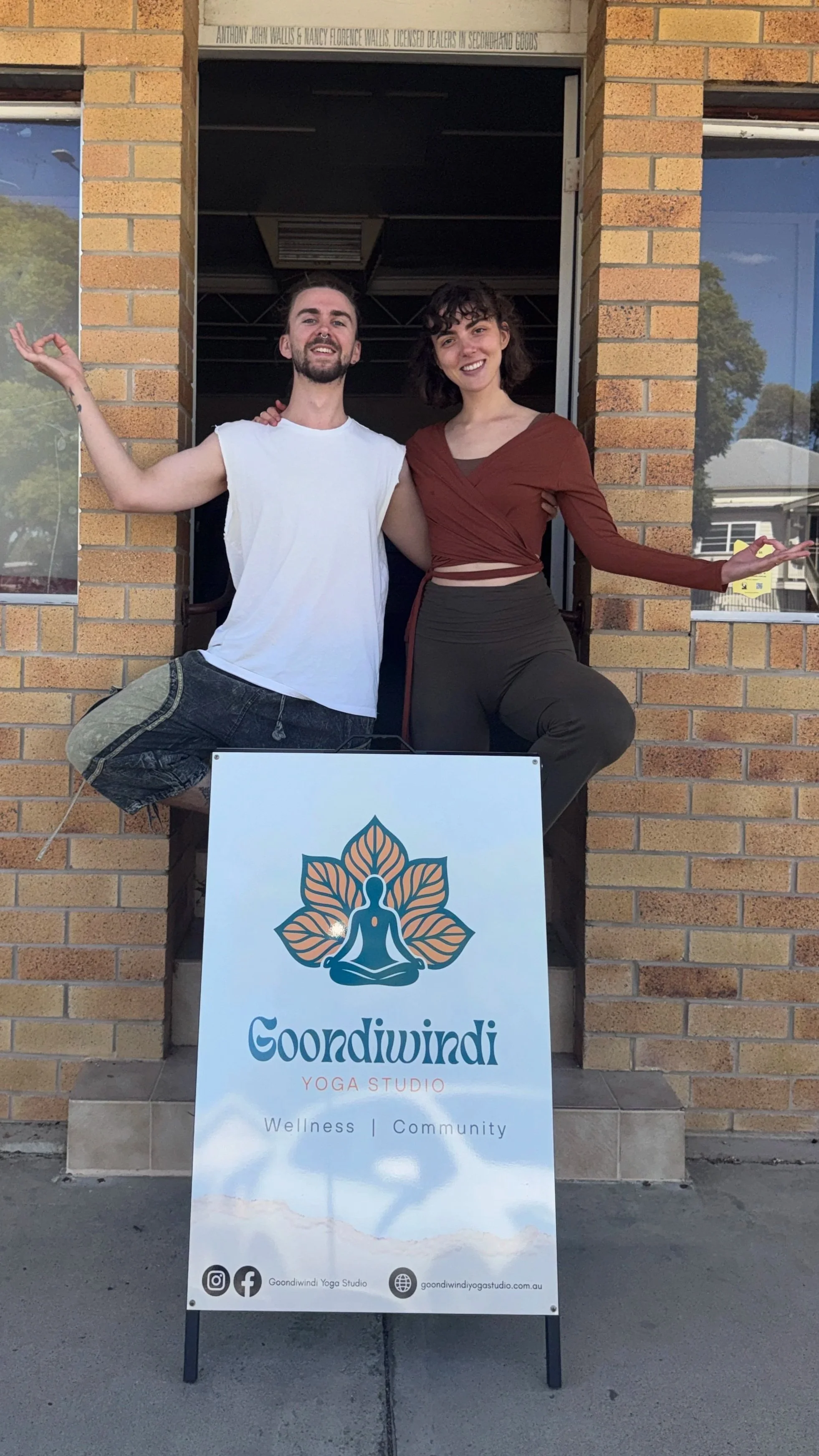 Two people standing inside the doorway of a yoga studio, smiling and posing with their arms around each other. In front of them is a sign that says 'Goindiwindi Yoga Studio' with the words 'Wellness' and 'Community' underneath, along with social media icons and the studio's website.