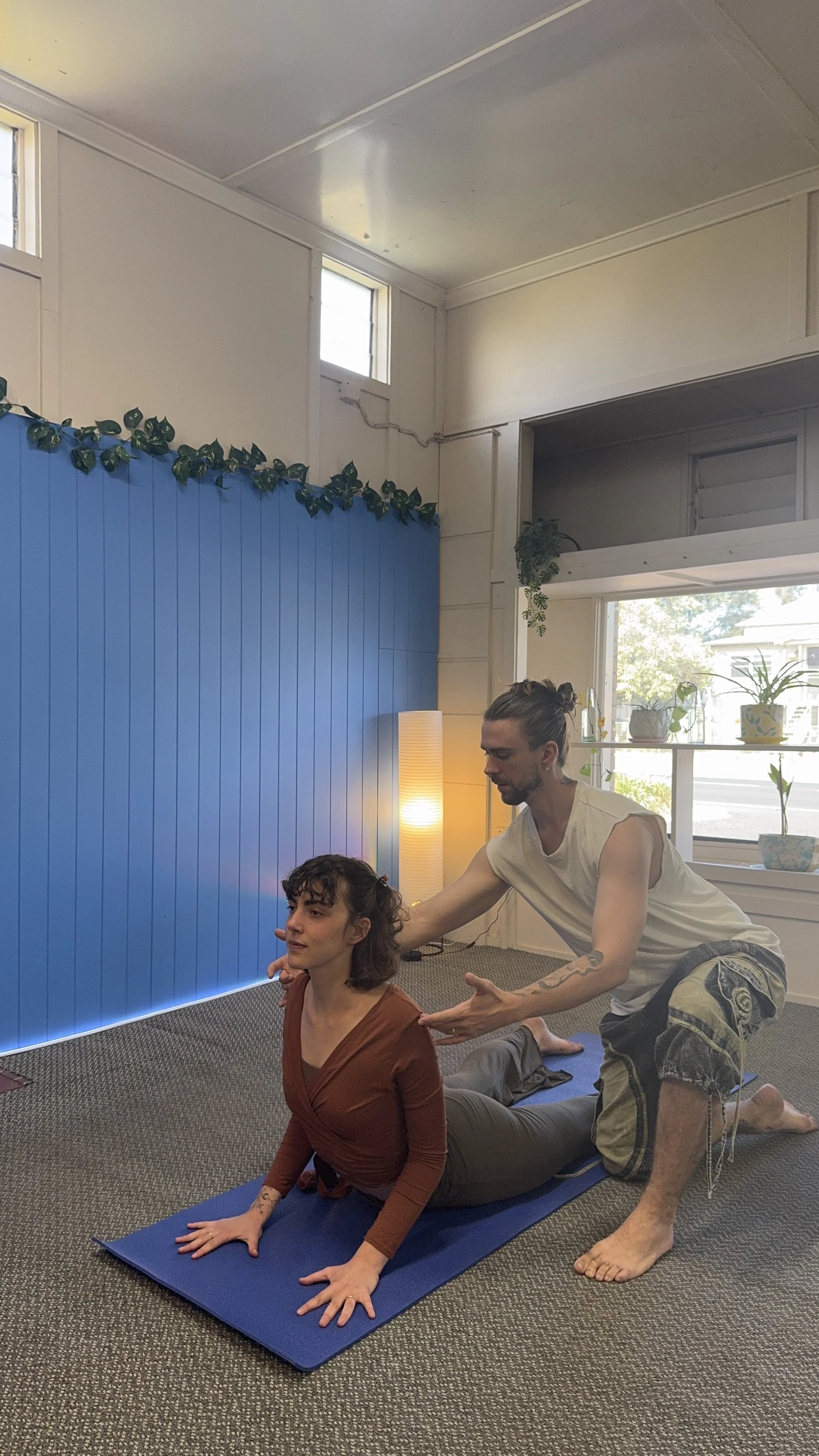 A man practicing yoga with a woman in a yoga pose in a room with natural light, blue wall, and potted plants.