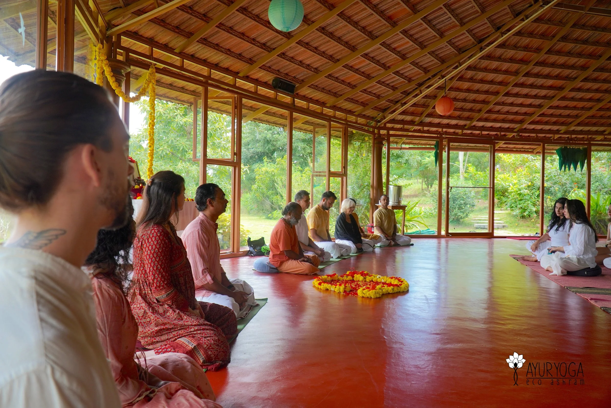 People seated in a circle during a yoga or meditation session inside a wooden-floored room with large open windows and green outdoor scenery.