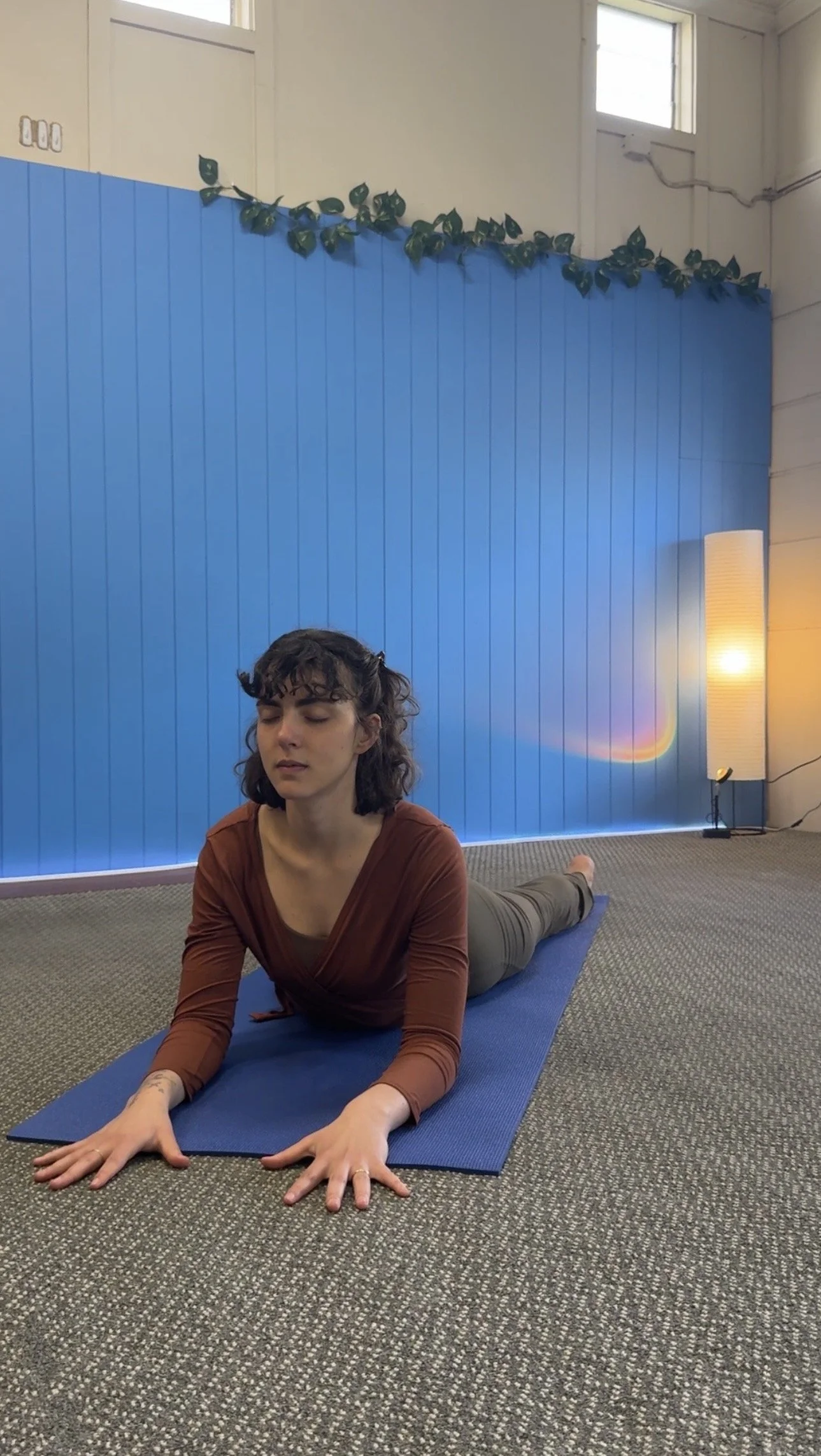 A woman with dark curly hair practicing yoga on a blue mat indoors, with a blue wall, a window, a tall lamp, and a patterned carpet in the background.