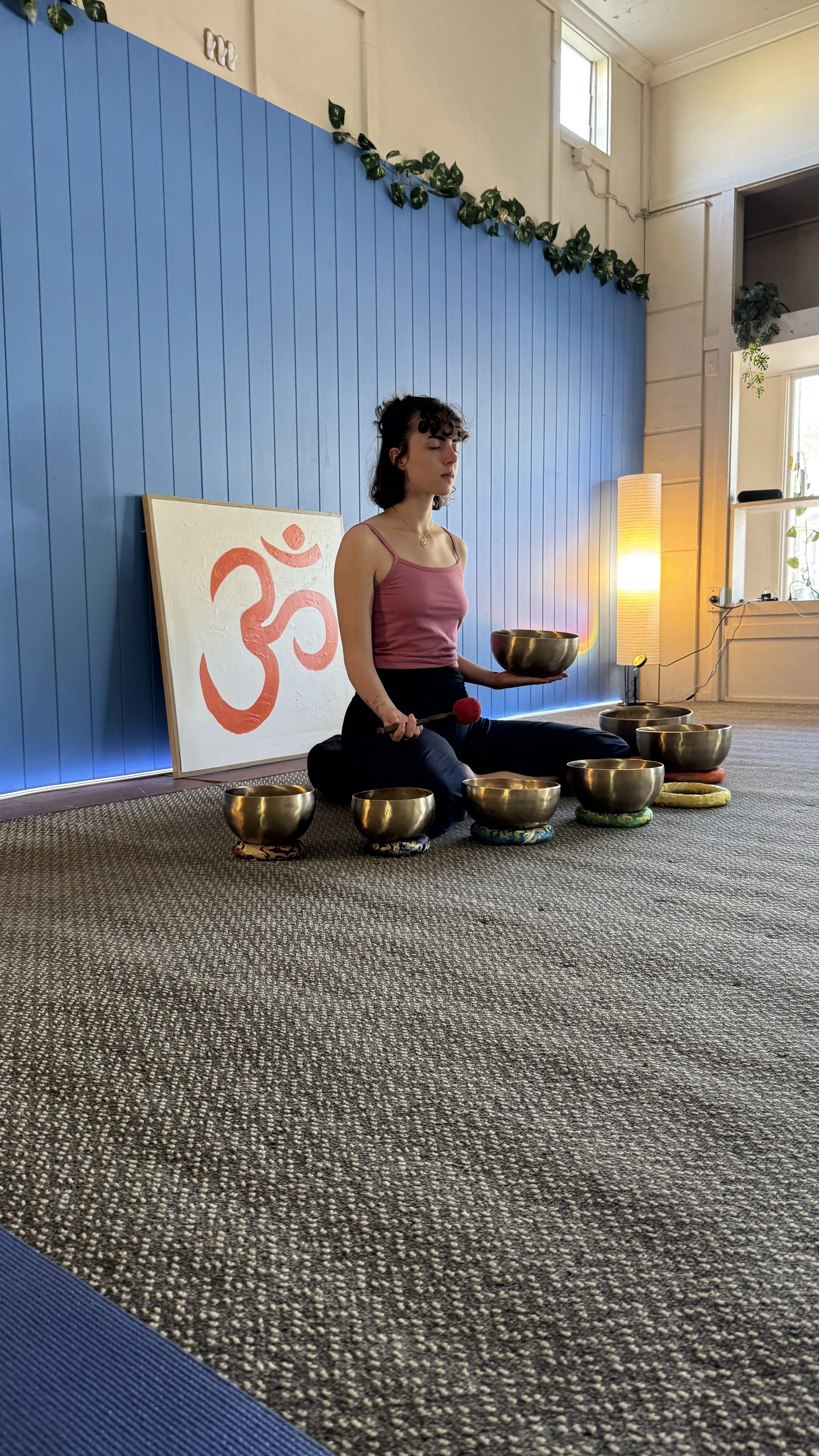 A woman sitting on the floor in a yoga or meditation pose, holding metal singing bowls with a mallet in a room with a blue wall, a painting with an Om symbol, and a tall lamp.