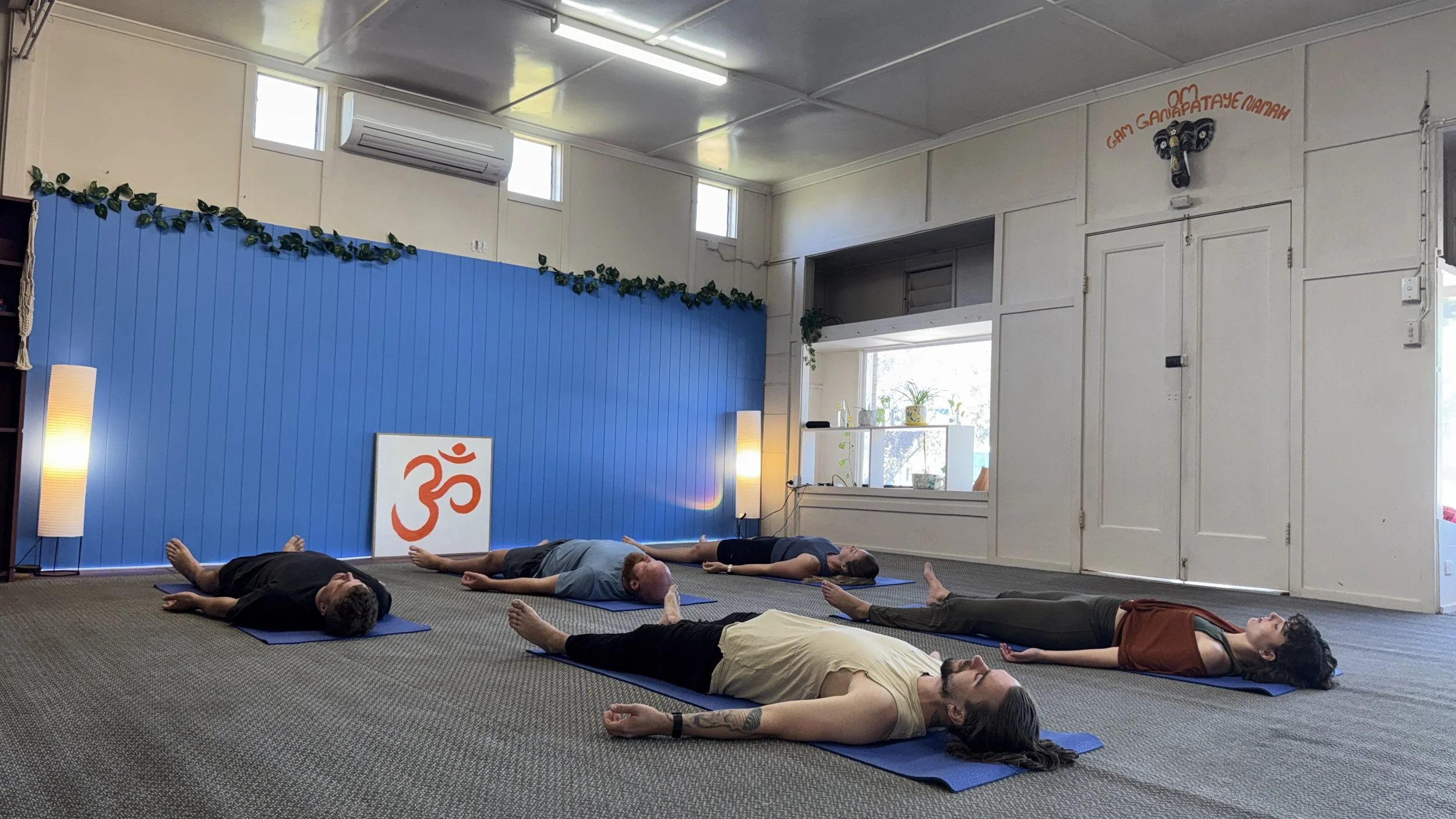 People lying on yoga mats in a yoga class, arranged in rows, practicing relaxation or meditation in a room with white walls, windows, and blue decor, including a sign with an orange Om symbol.