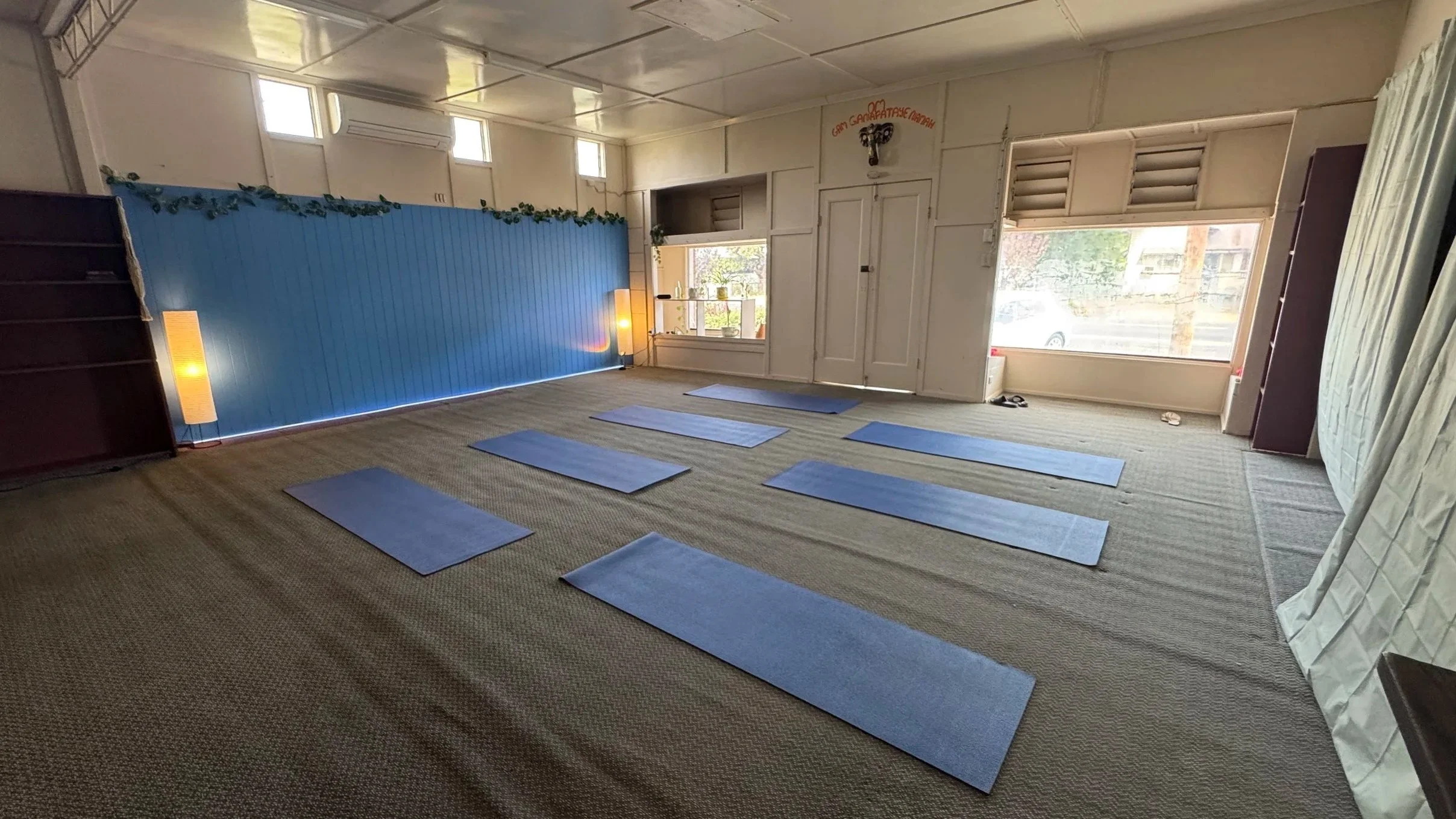 Indoor yoga room with blue yoga mats on a beige carpet. Windows let in natural light, and there is a blue partition with green decorative leaves. A sign with red text is visible on the wall near the ceiling.