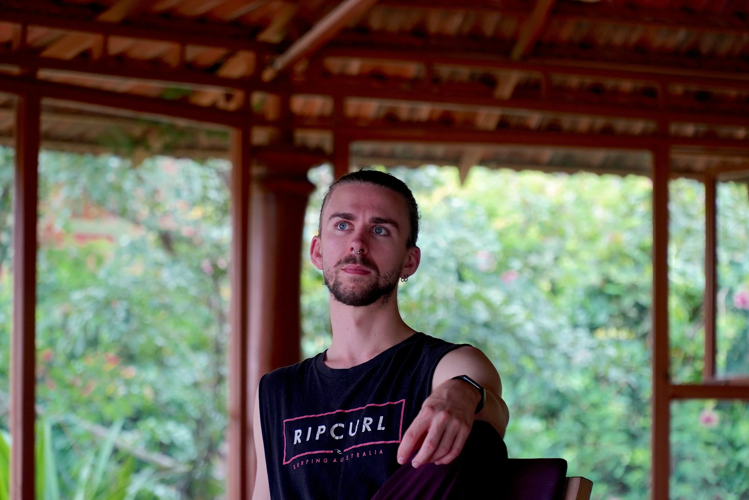 A person with a septum piercing and earrings, wearing a black Rip Curl T-shirt, stands on a porch with a wooden roof, greenery in the background, looking to the side.