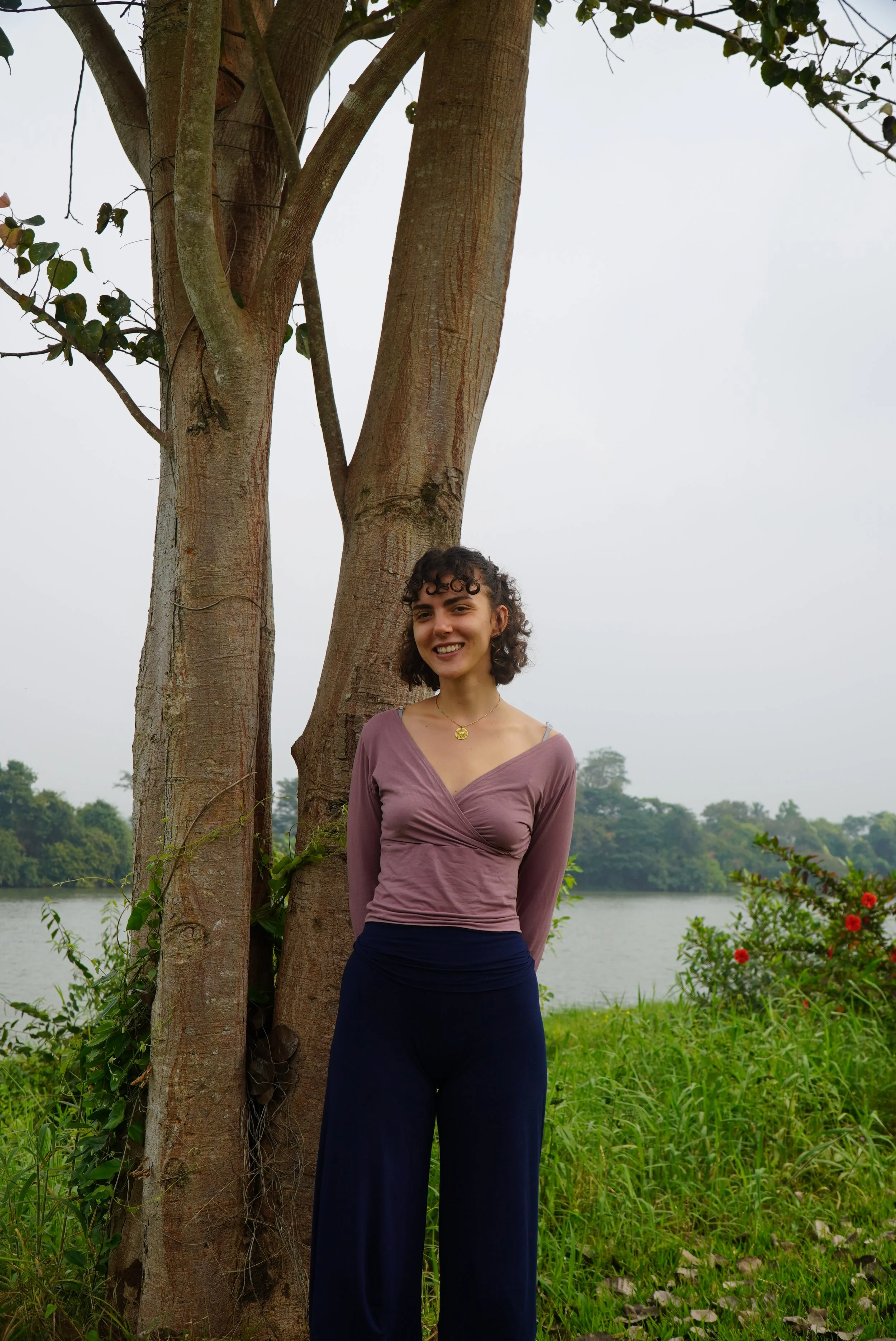 A woman with curly hair wearing a mauve long-sleeve top and dark pants standing beside a tree near a river, with lush greenery and a cloudy sky in the background.