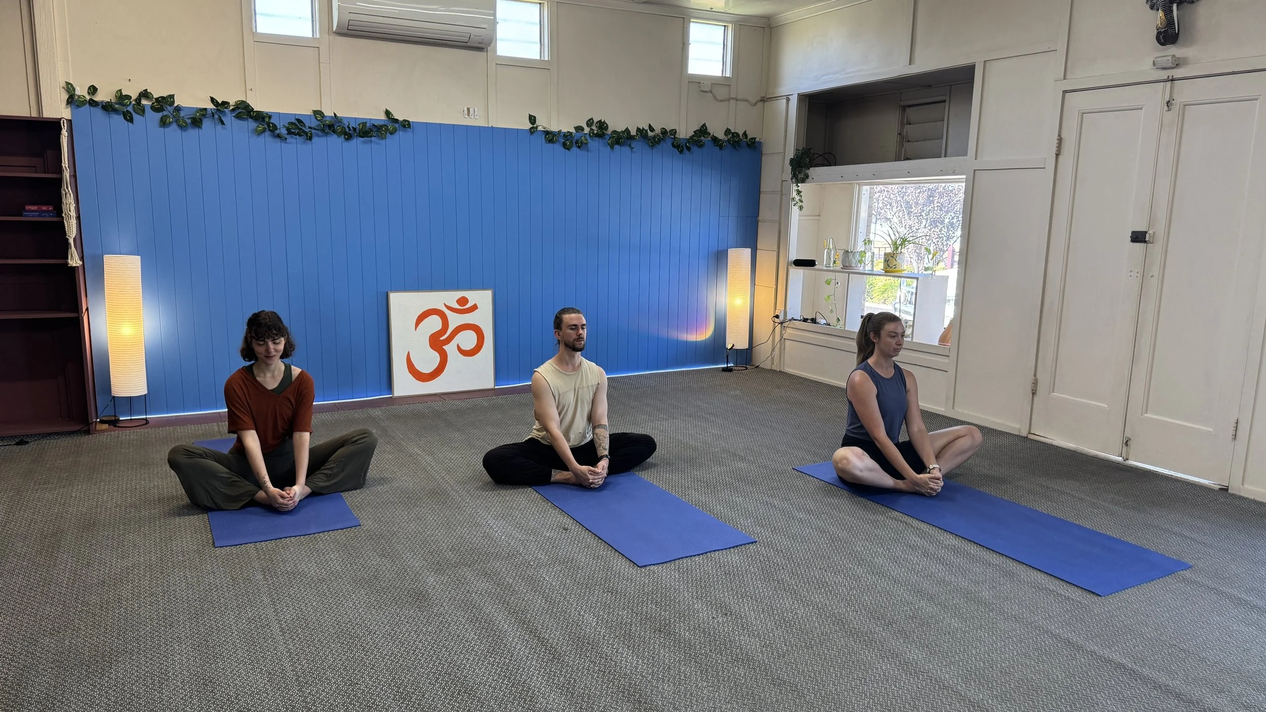 Three people in a yoga studio sitting cross-legged on mats with closed eyes, participating in a meditation session. The studio has a blue wall with a decorative green plant border, a white framed Om symbol, and large windows letting in natural light.