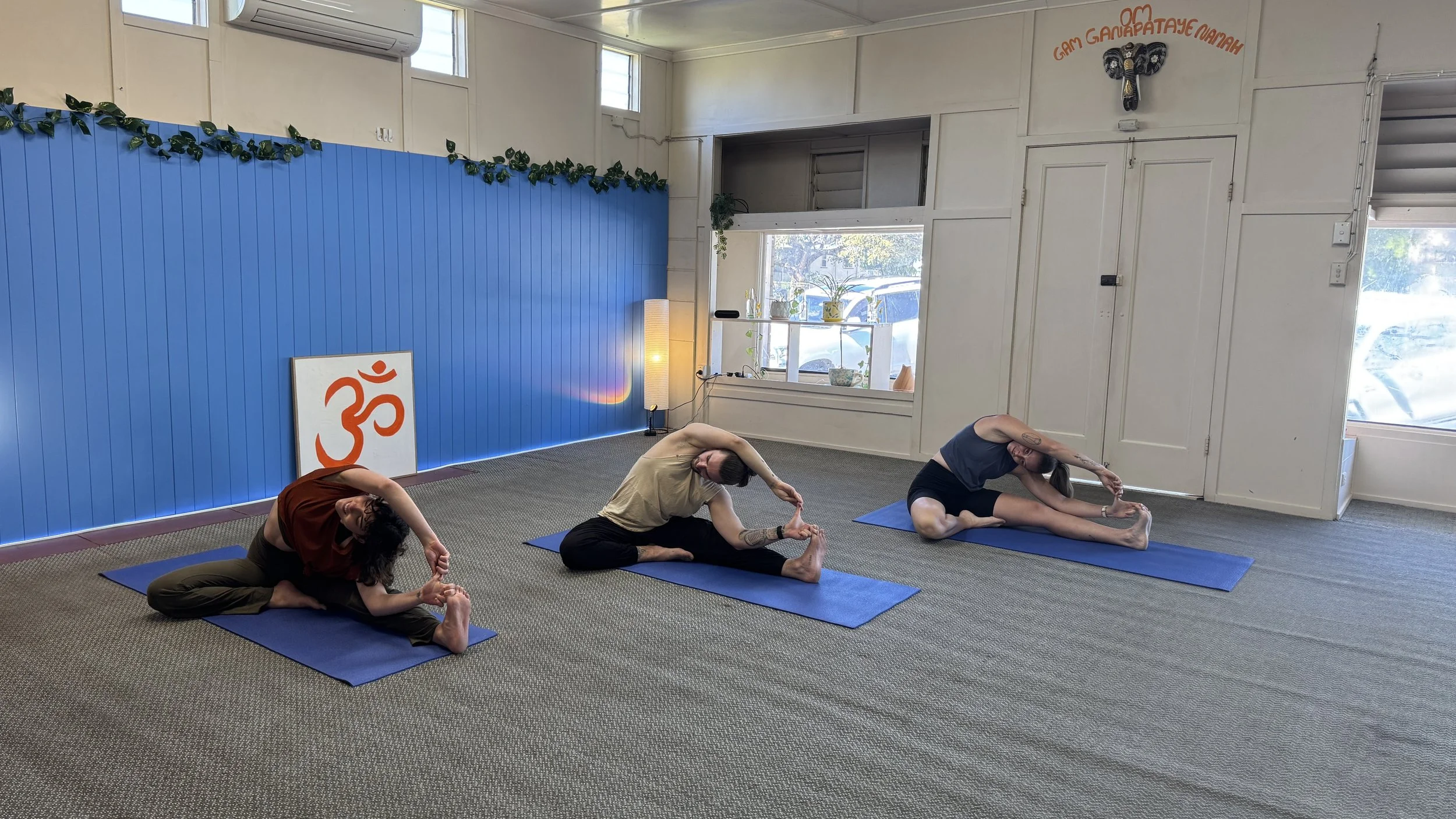 Three people practicing yoga in a room with blue wall paneling, a sign with an orange symbol, plants on the window sill, and a sign with orange text and a decorative elephant. They are seated on blue mats, stretching their arms over their heads and t