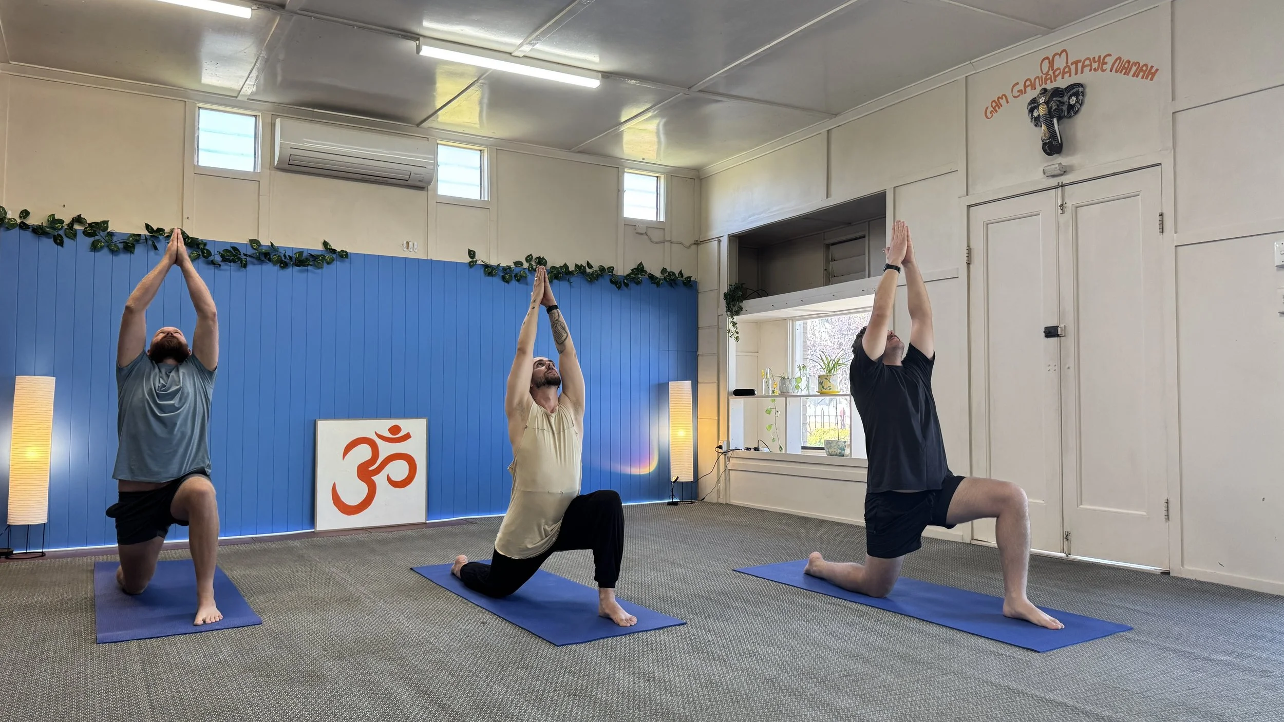 Three men practicing yoga in a room with blue and white walls, a large orange Om symbol, and decorative plants. All are in a kneeling yoga pose with hands raised above their heads, on yoga mats.