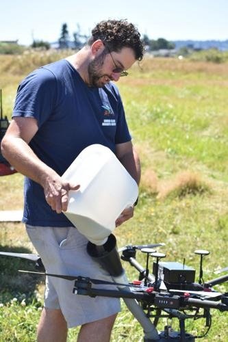 A man with curly hair and glasses is standing outdoors in a grassy field, holding a white container and operating a large drone with multiple rotors.
