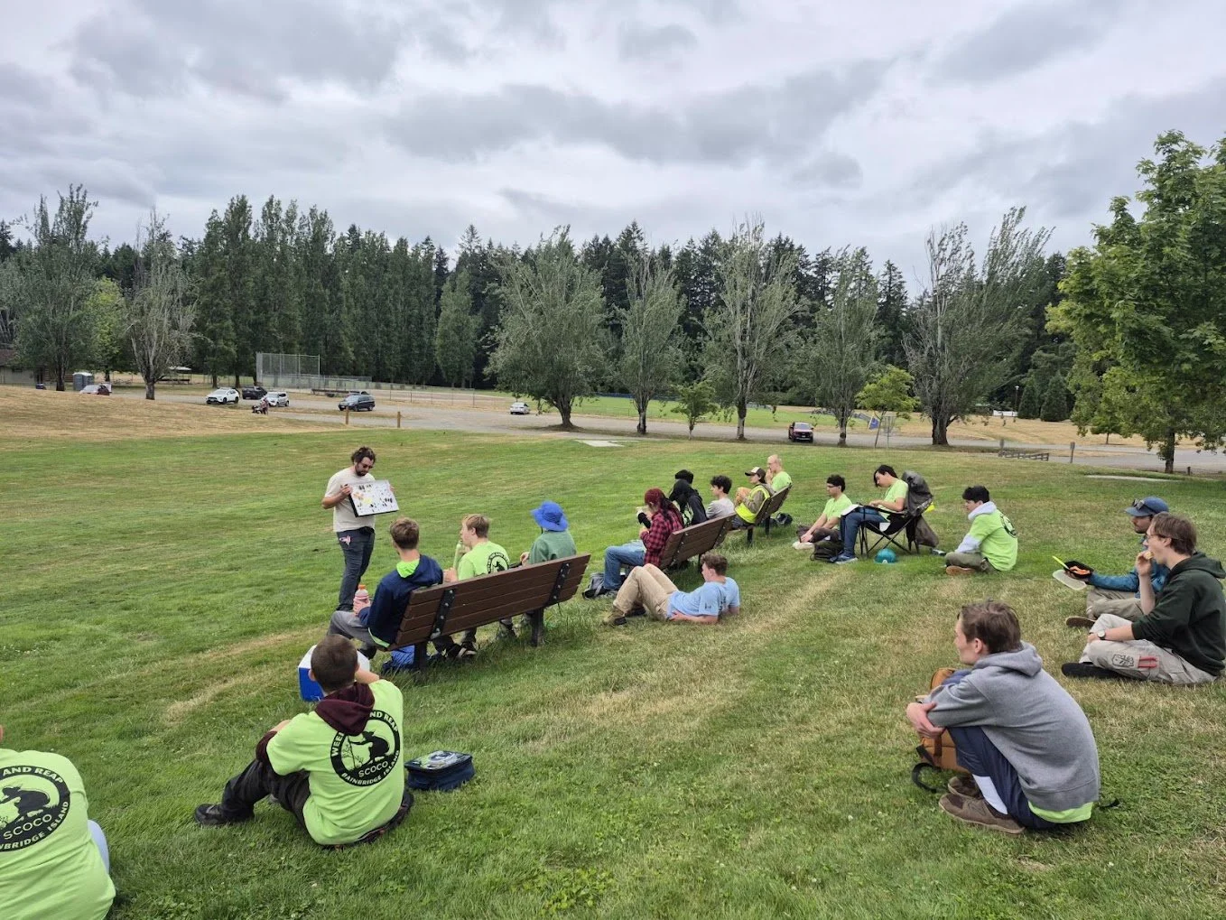 Group of people sitting on a grassy field listening to a person standing and holding a large poster or board, outdoors on a cloudy day, with trees and a parking lot in the background.